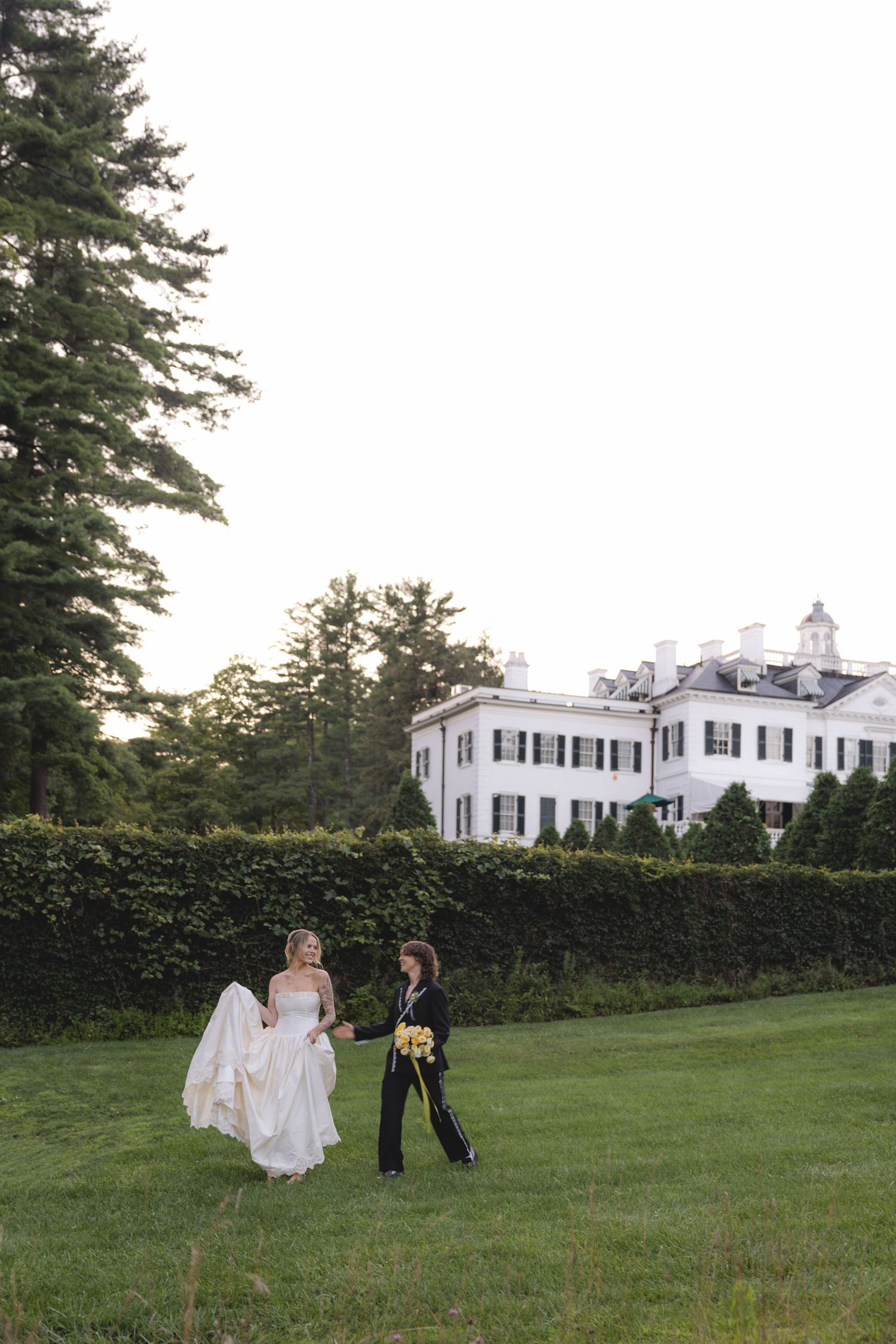 Same-sex couple in wedding attire walks on green lawn before white colonial mansion with black shutters.