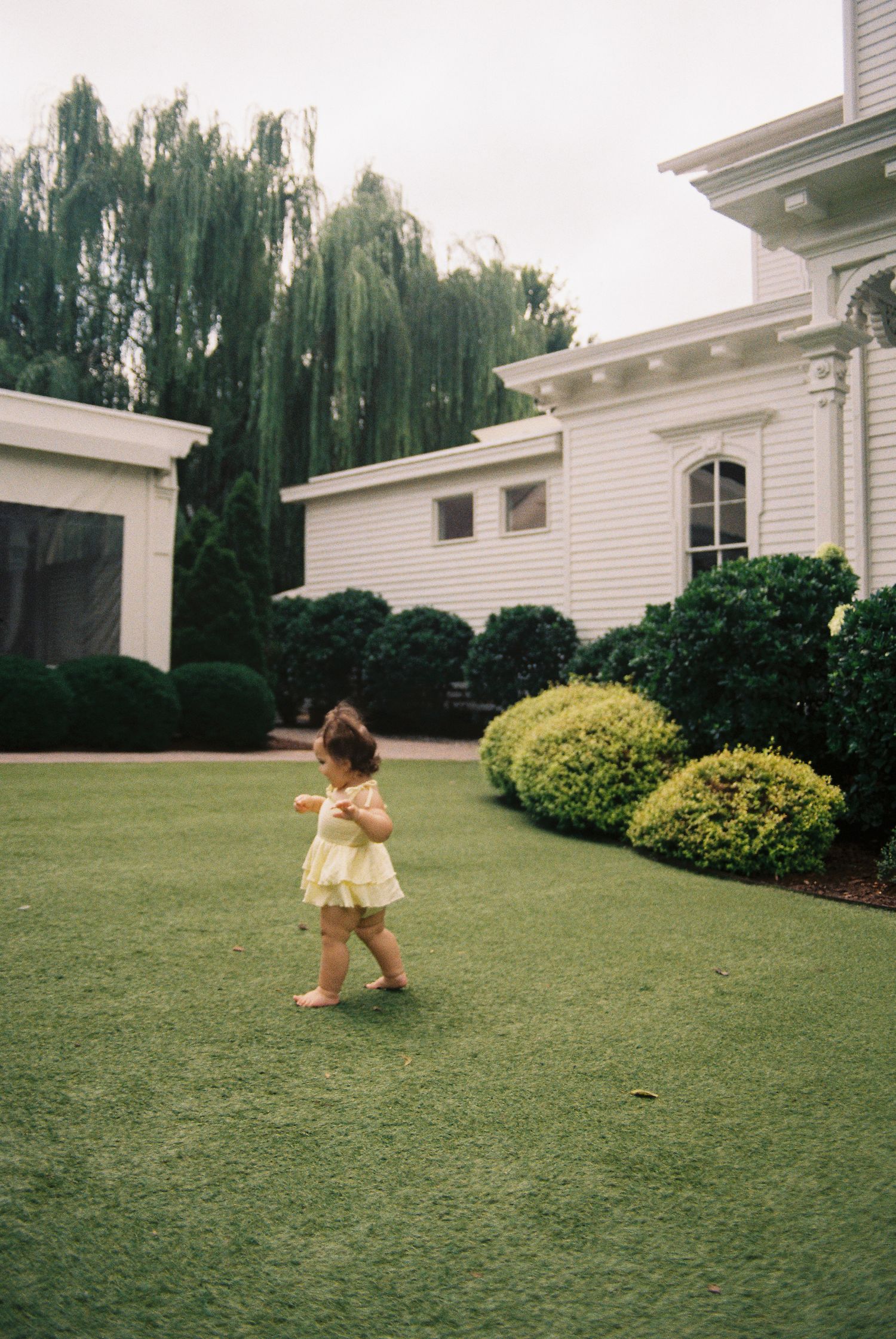 Toddler girl in yellow dress playing on lush green lawn beside white colonial home with manicured shrubs.