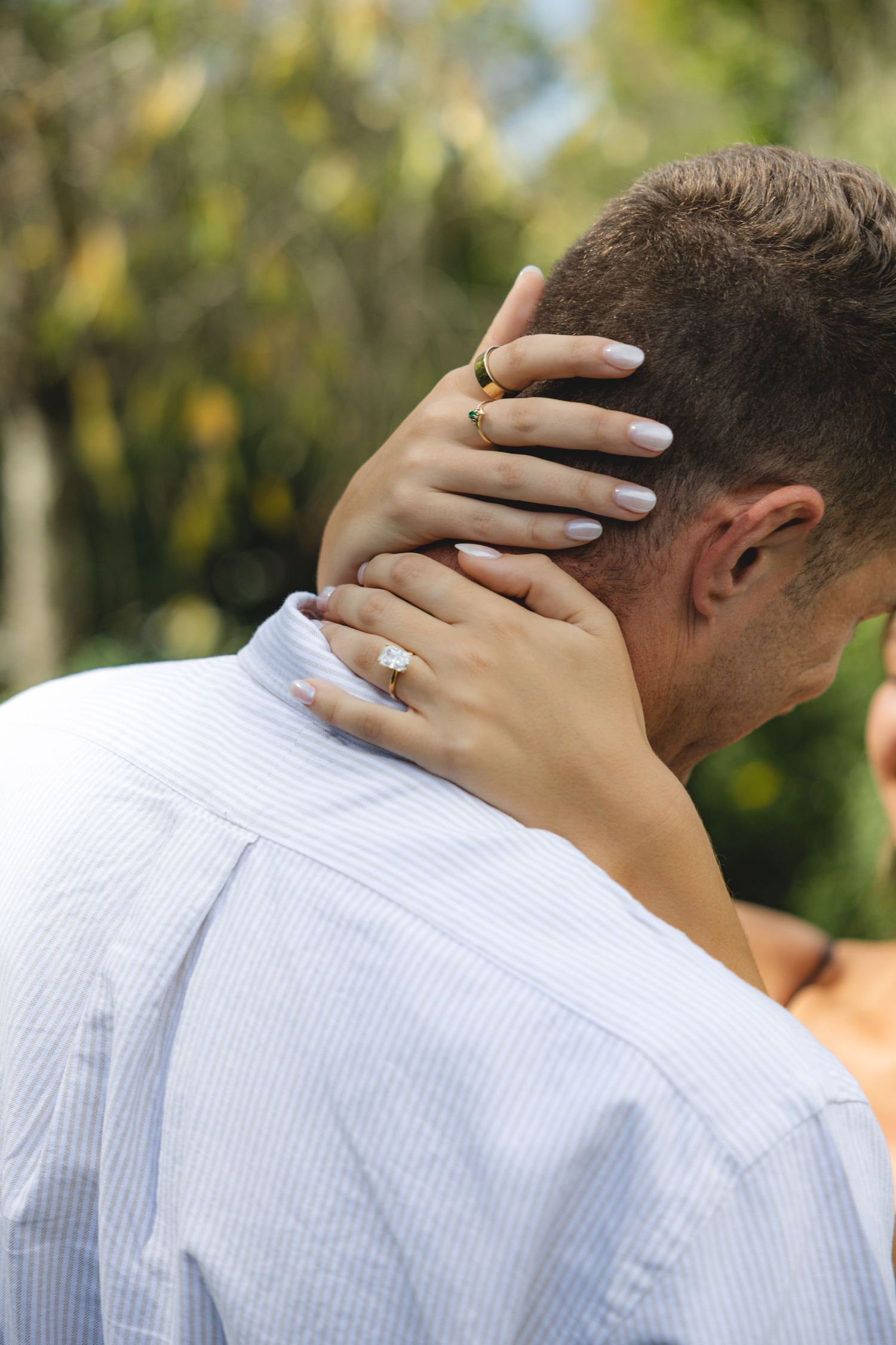 Woman's hand with engagement ring resting on man's neck in romantic outdoor setting.
