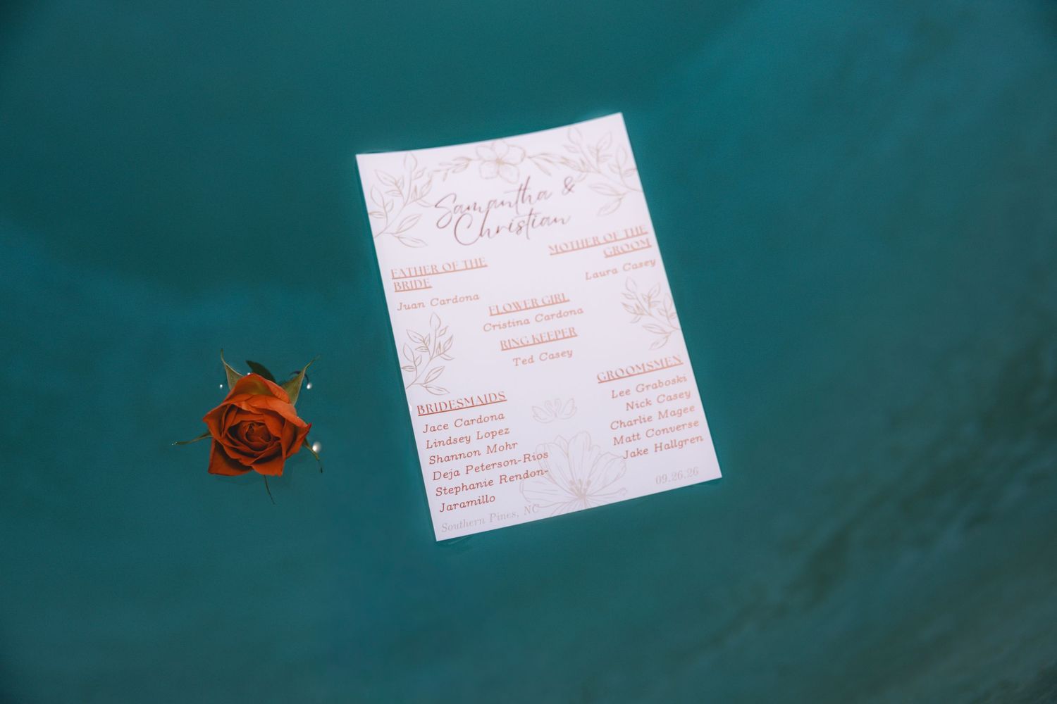 Wedding program and red rose on teal water surface at an outdoor ceremony.