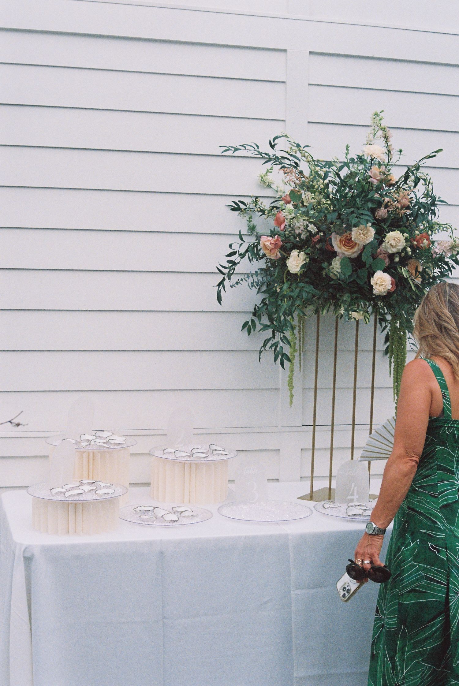Woman in green dress viewing wedding escort card table with floral arrangement on gold stand.
