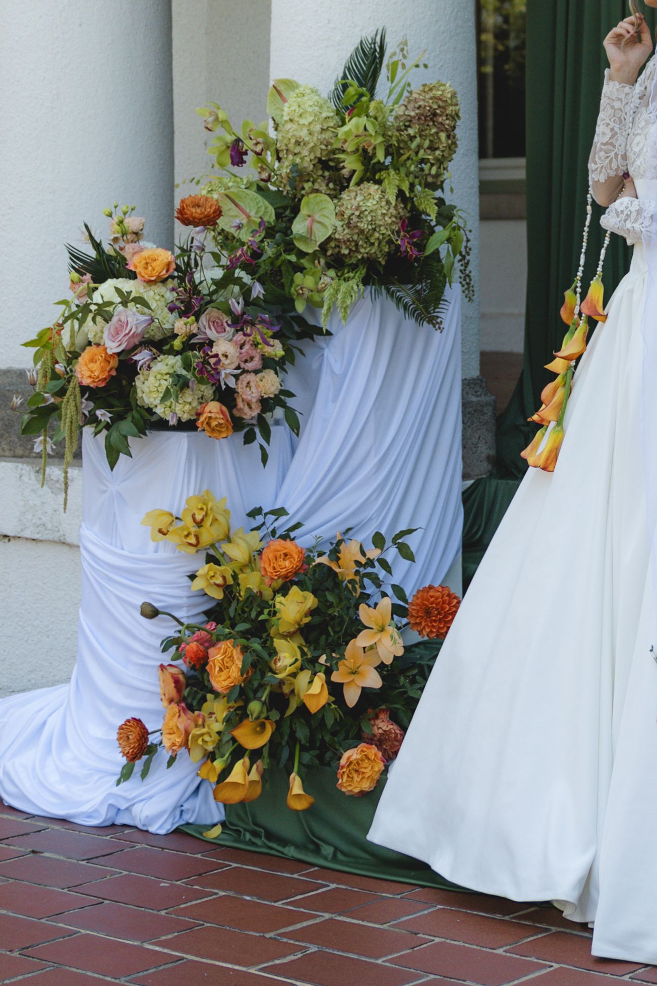 Bride in white gown stands beside lush orange and yellow floral arrangements on draped white pedestals at wedding venue.