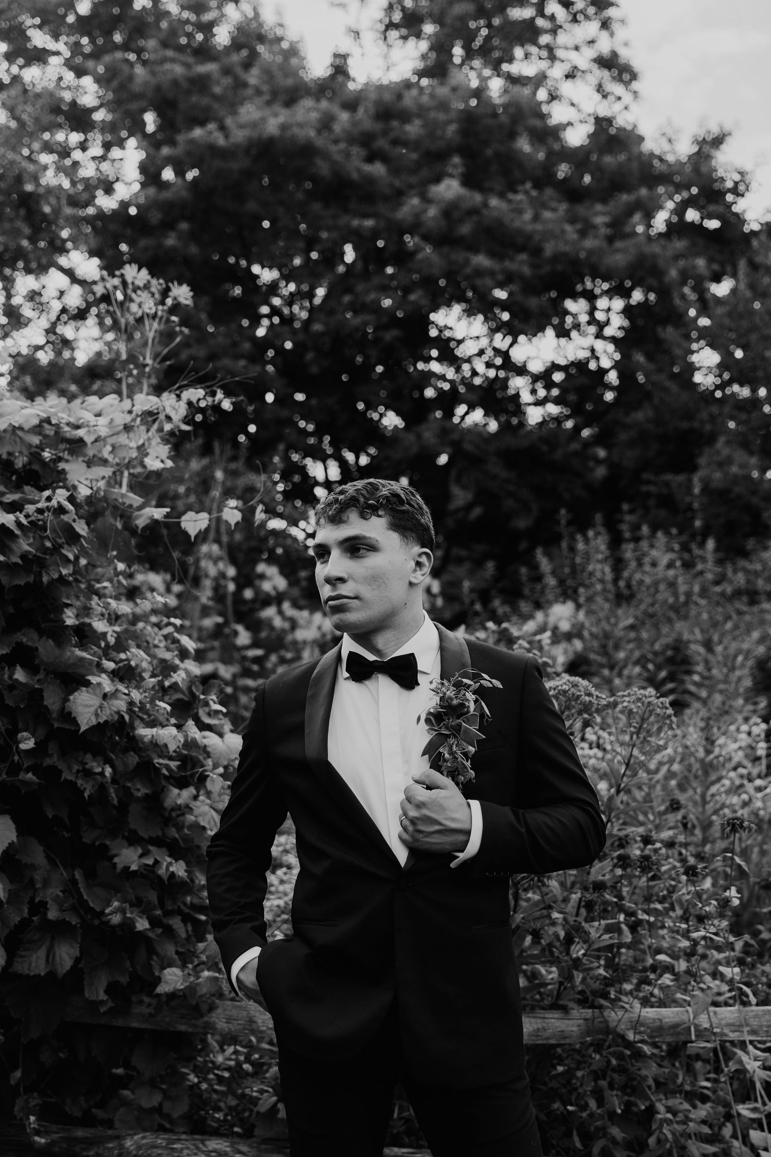 A groom in a black tuxedo with bow tie and boutonniere poses outdoors among lush greenery in a black and white photo.