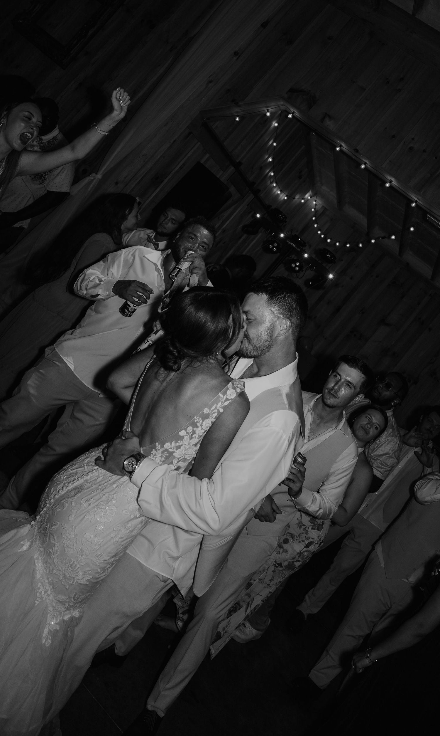 Black and white photo of a bride and groom sharing a first dance surrounded by guests at a rustic wedding reception.
