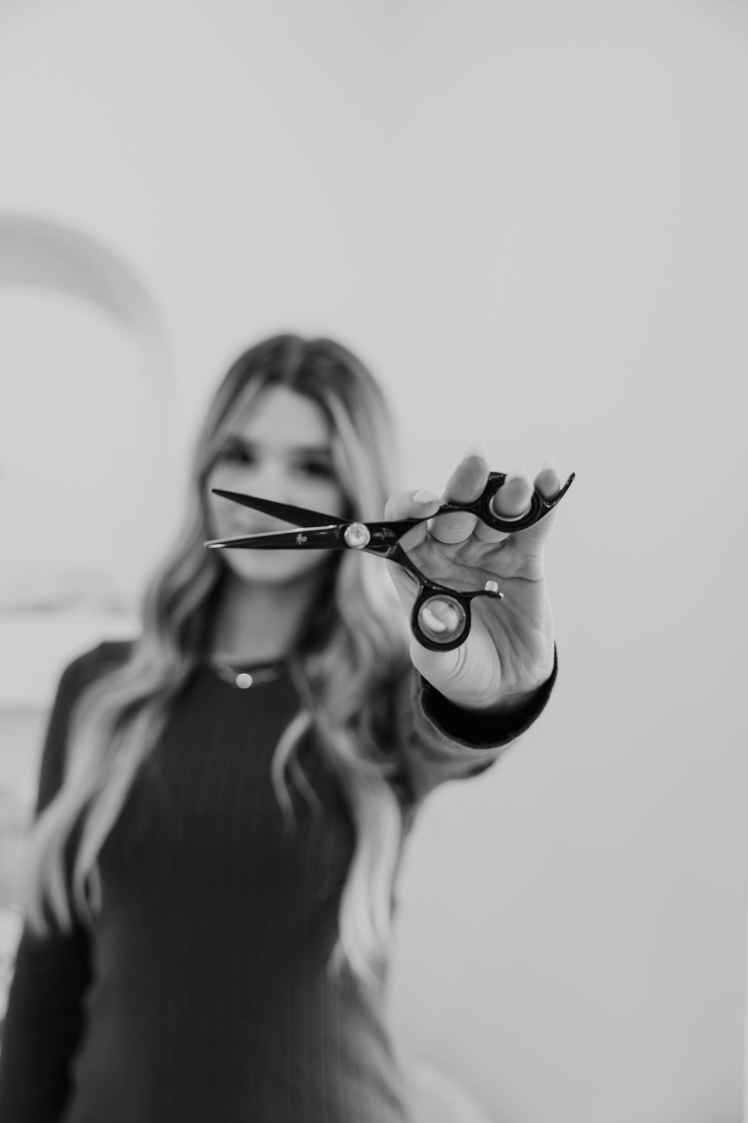 Black and white photo of a female hairstylist holding professional scissors toward the camera in a salon.