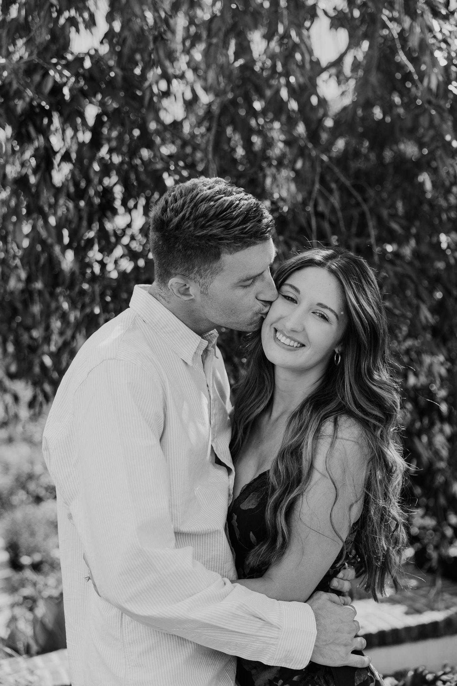 Black and white photo of a couple embracing outdoors, man kissing woman on cheek as she smiles, surrounded by lush greenery.