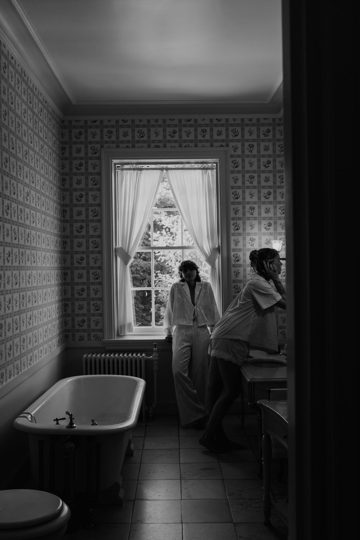 Black and white photo of two people in a vintage bathroom with clawfoot tub, patterned wallpaper, and window with curtains.