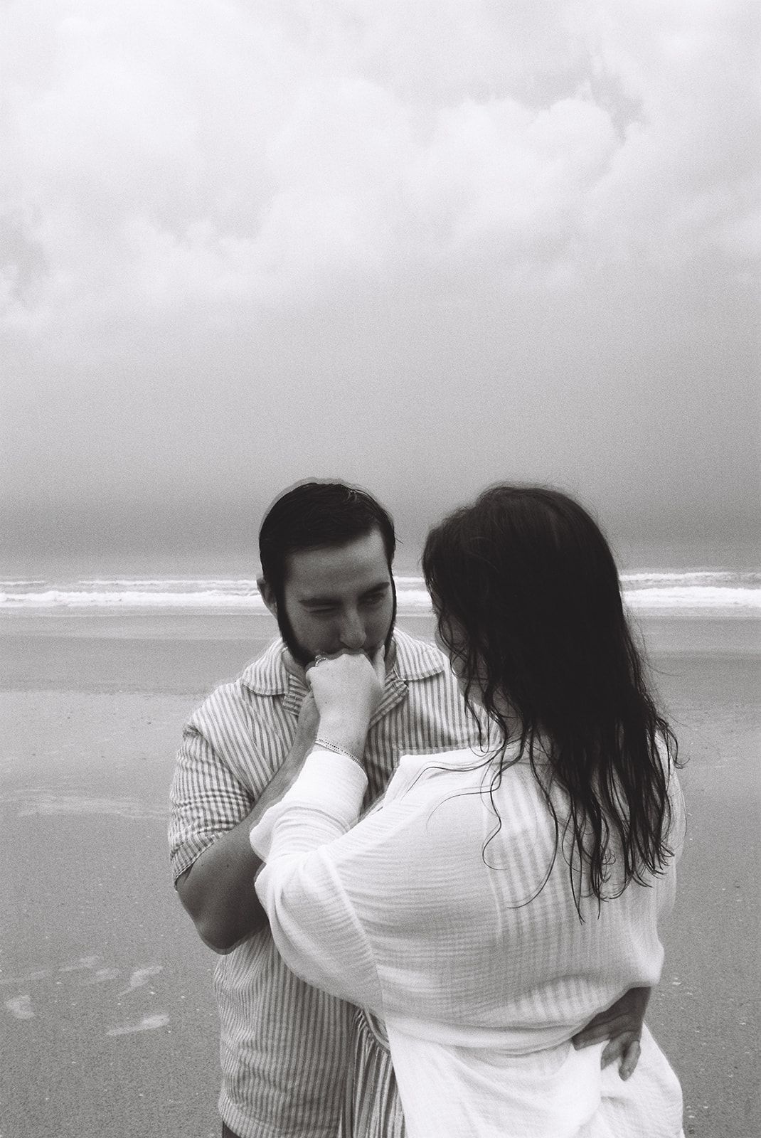 Black and white photo of a couple embracing on a cloudy beach, the man kissing the woman's hand tenderly.
