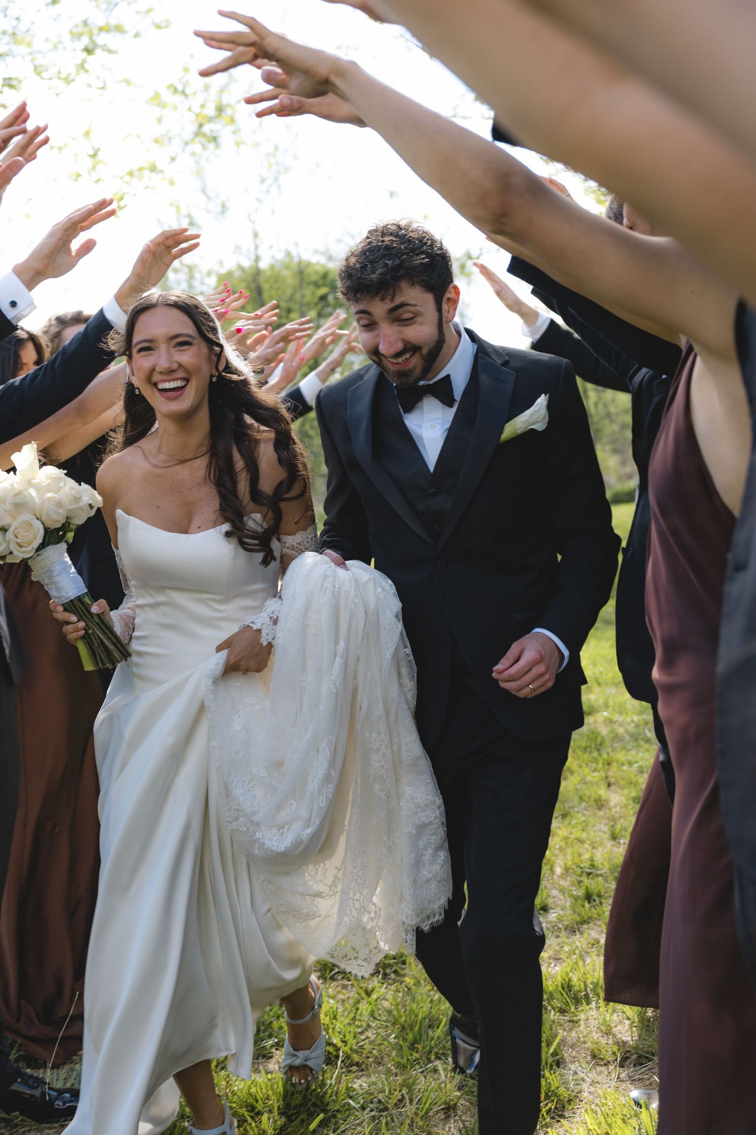 Laughing bride and groom walk through tunnel of raised hands formed by wedding party outdoors.