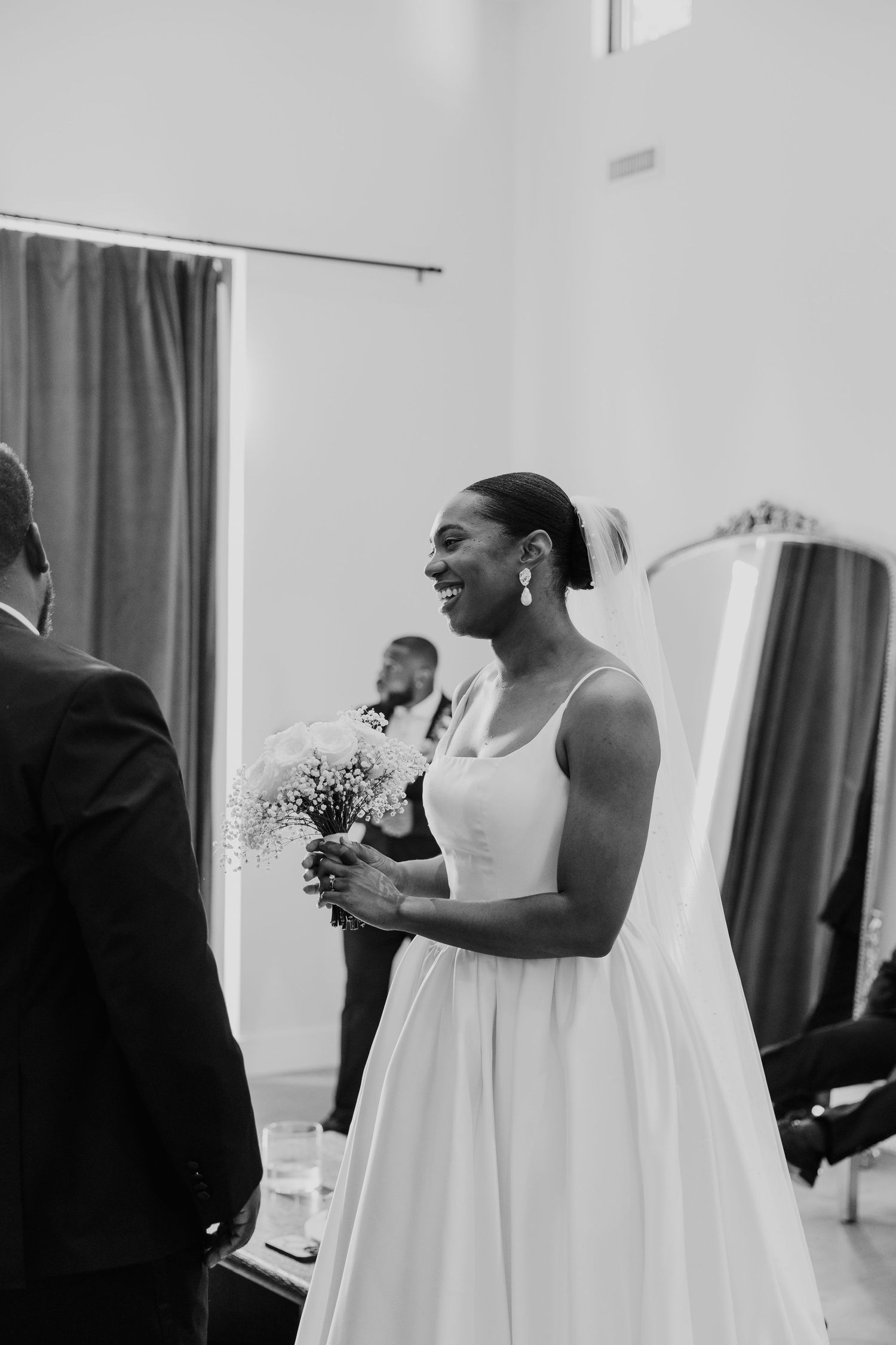 Black and white photo of radiant bride holding bouquet and smiling in bridal suite before ceremony.