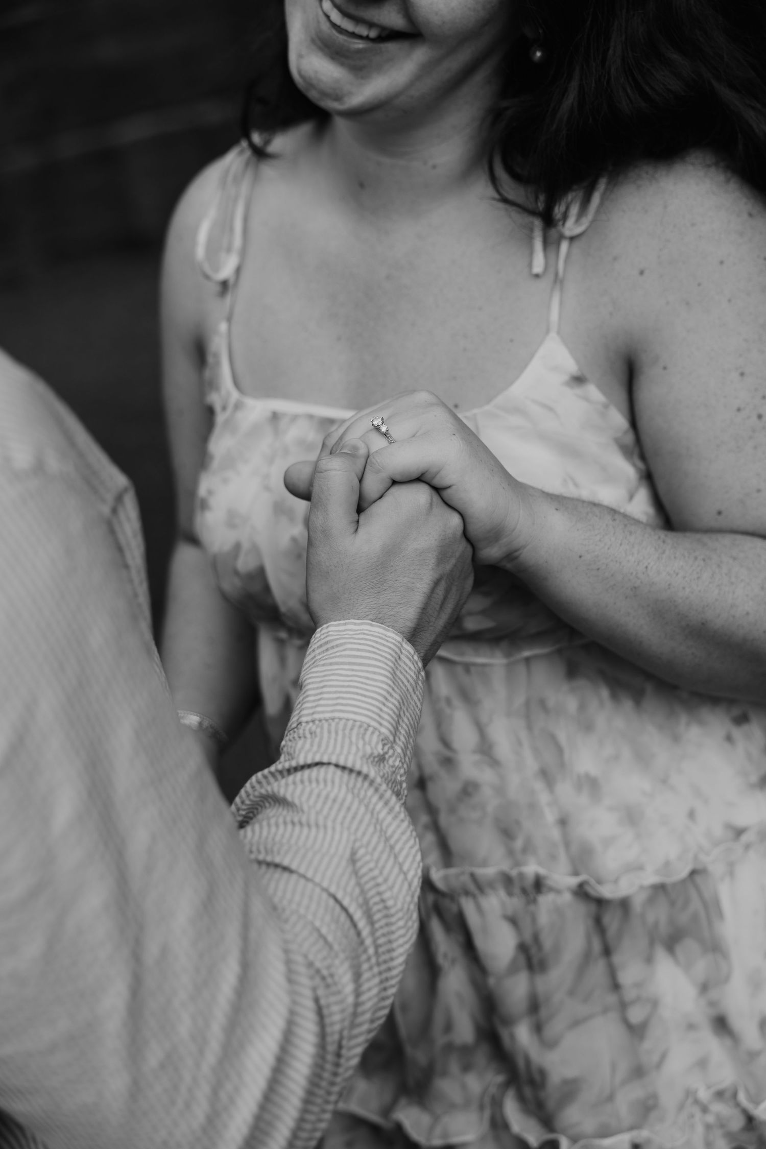 Black and white photo of a couple holding hands, woman wearing engagement ring and floral dress, smiling joyfully.