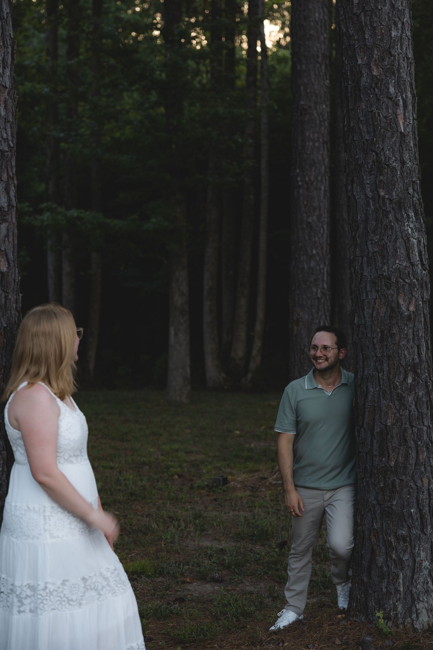 Couple sharing a romantic moment in a forest, woman in white dress and man in green polo leaning against pine tree.