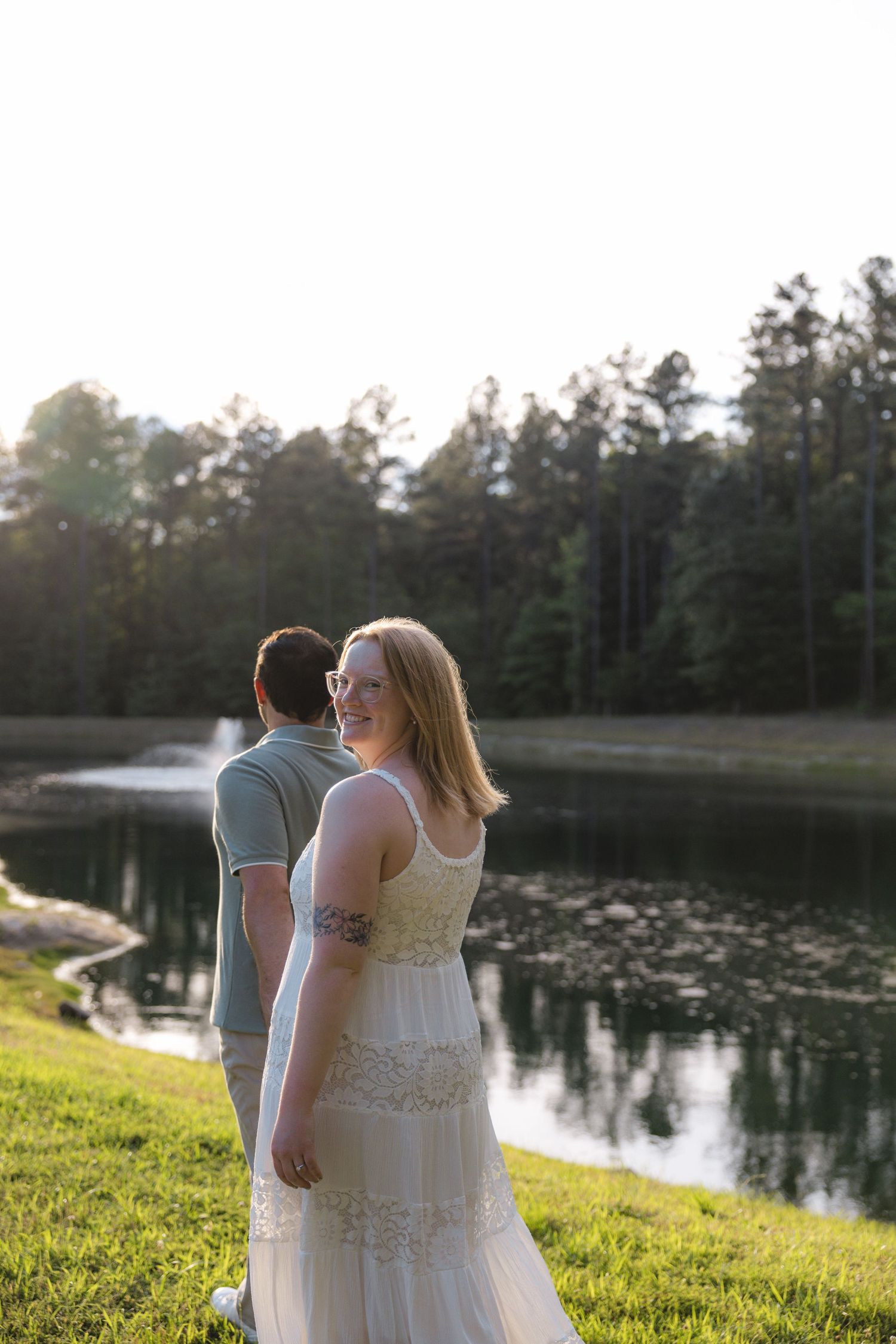 Bride in white lace gown smiles by a sunlit pond with groom in gray shirt, surrounded by tall pine trees.