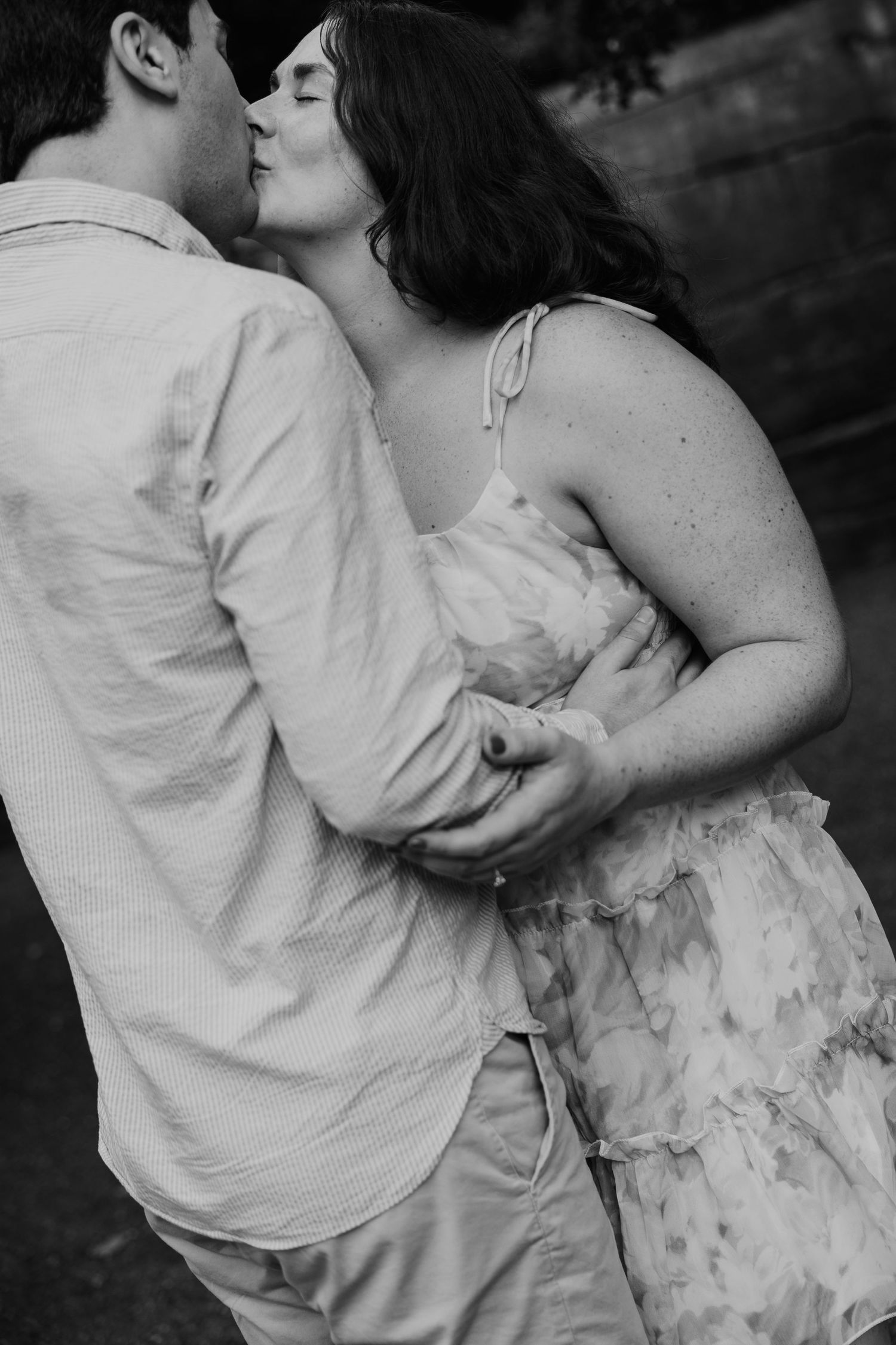 Black and white photo of a couple embracing and kissing outdoors, woman in floral dress, man in button-up shirt.