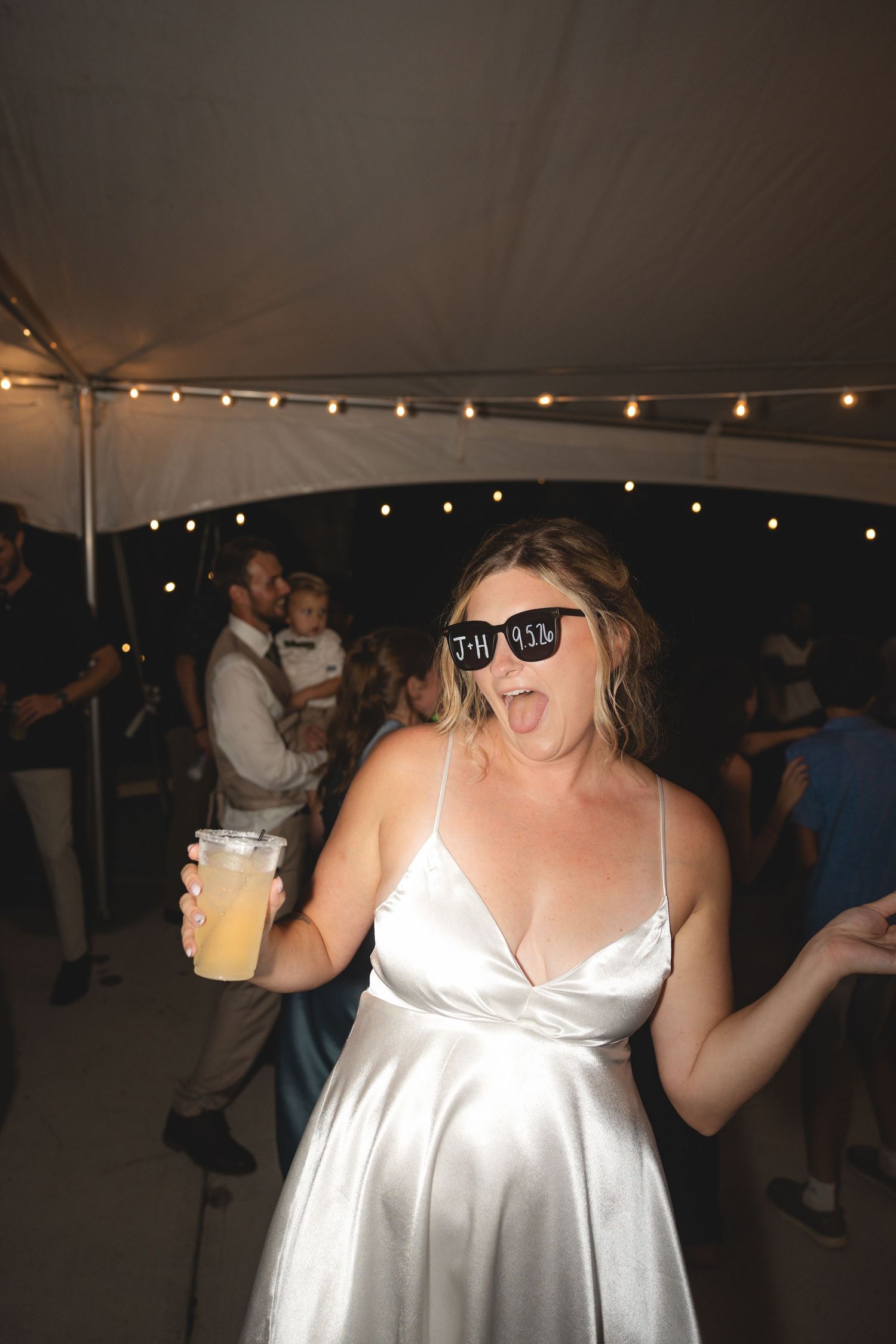 Bride in white satin dress and novelty sunglasses dancing with a drink at an outdoor wedding reception.