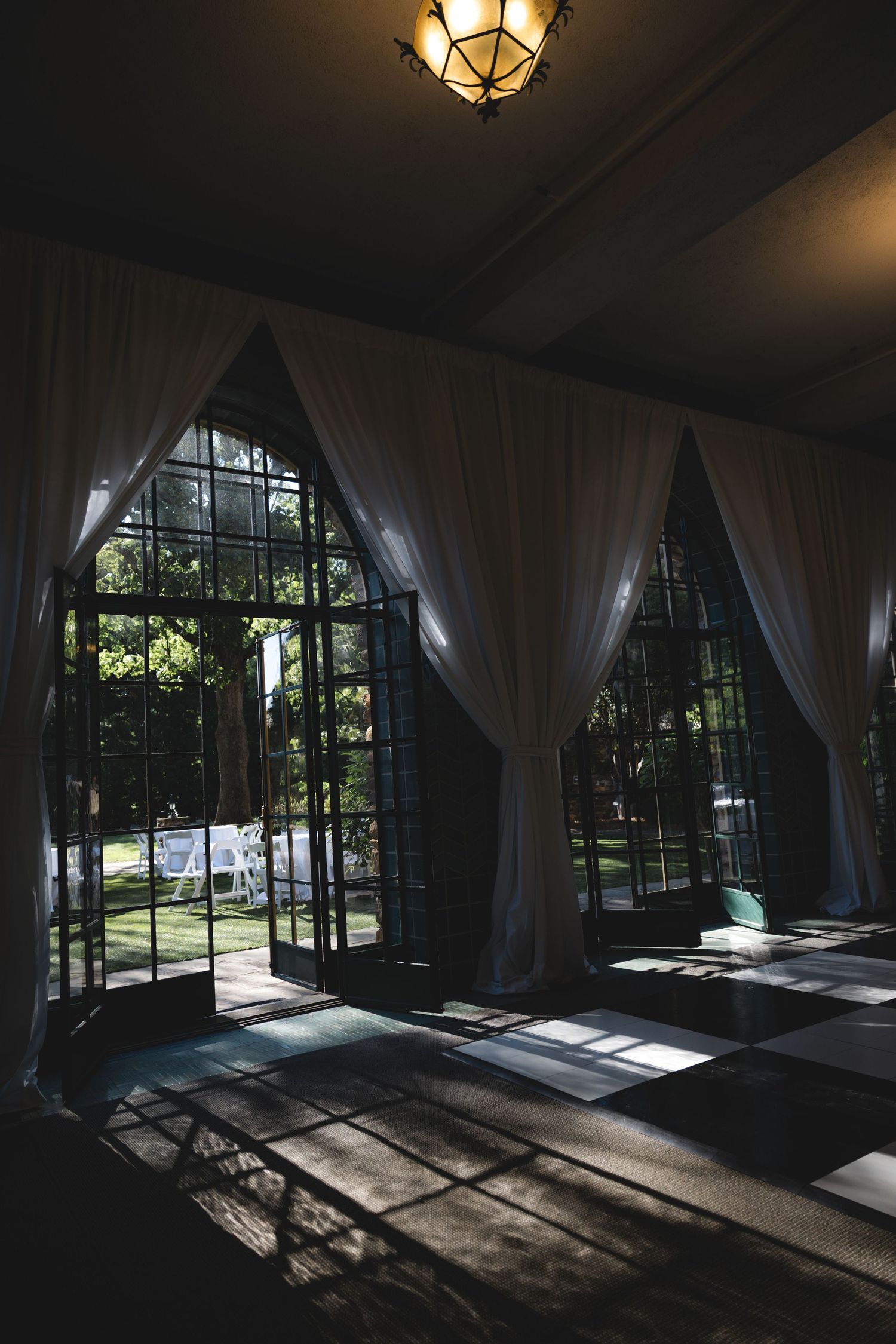 Elegant venue interior with white draped curtains, arched black-framed windows, and sunlight casting geometric shadows on the floor.