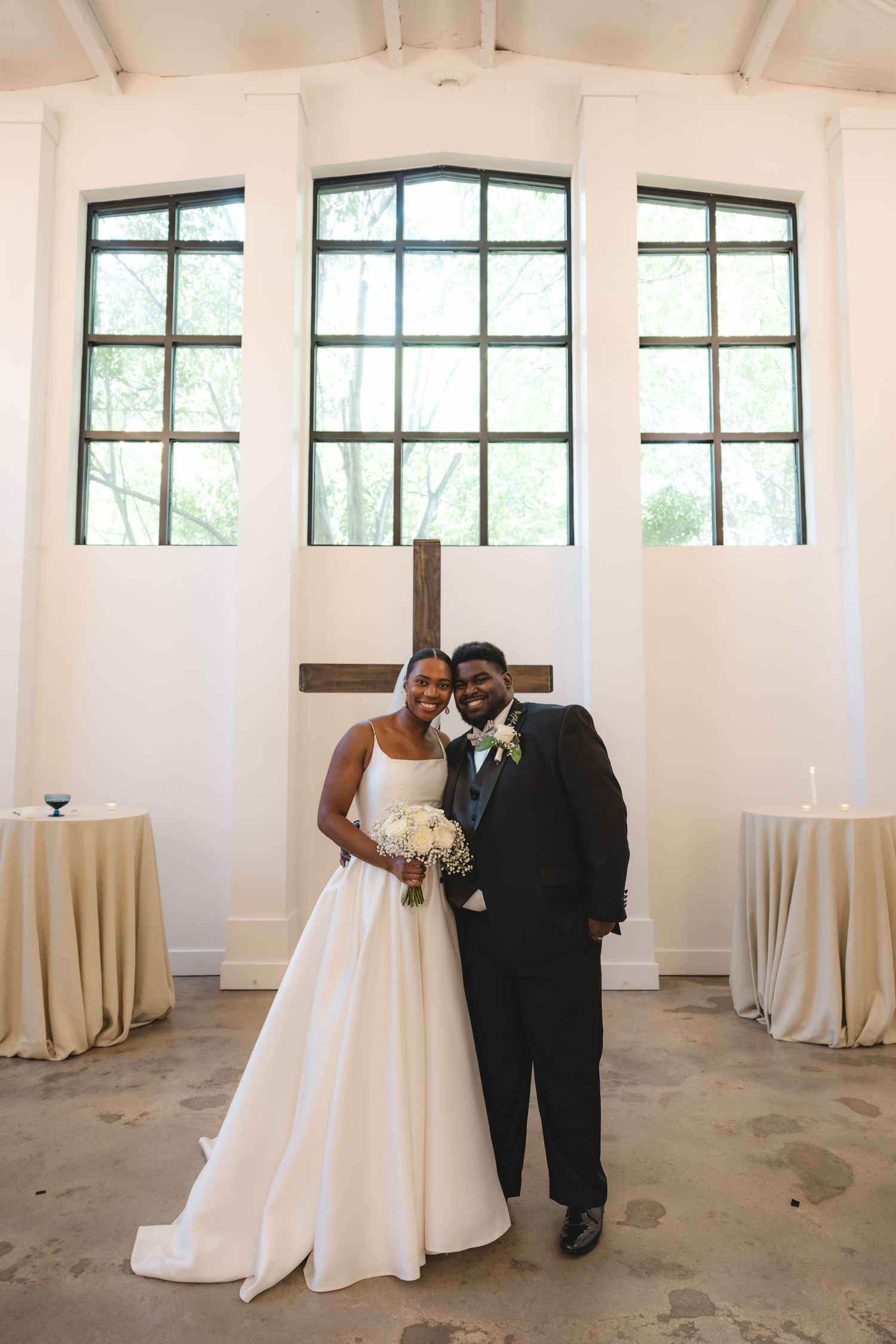 Bride and groom posing together in a white chapel with large windows and a wooden cross after their wedding ceremony.