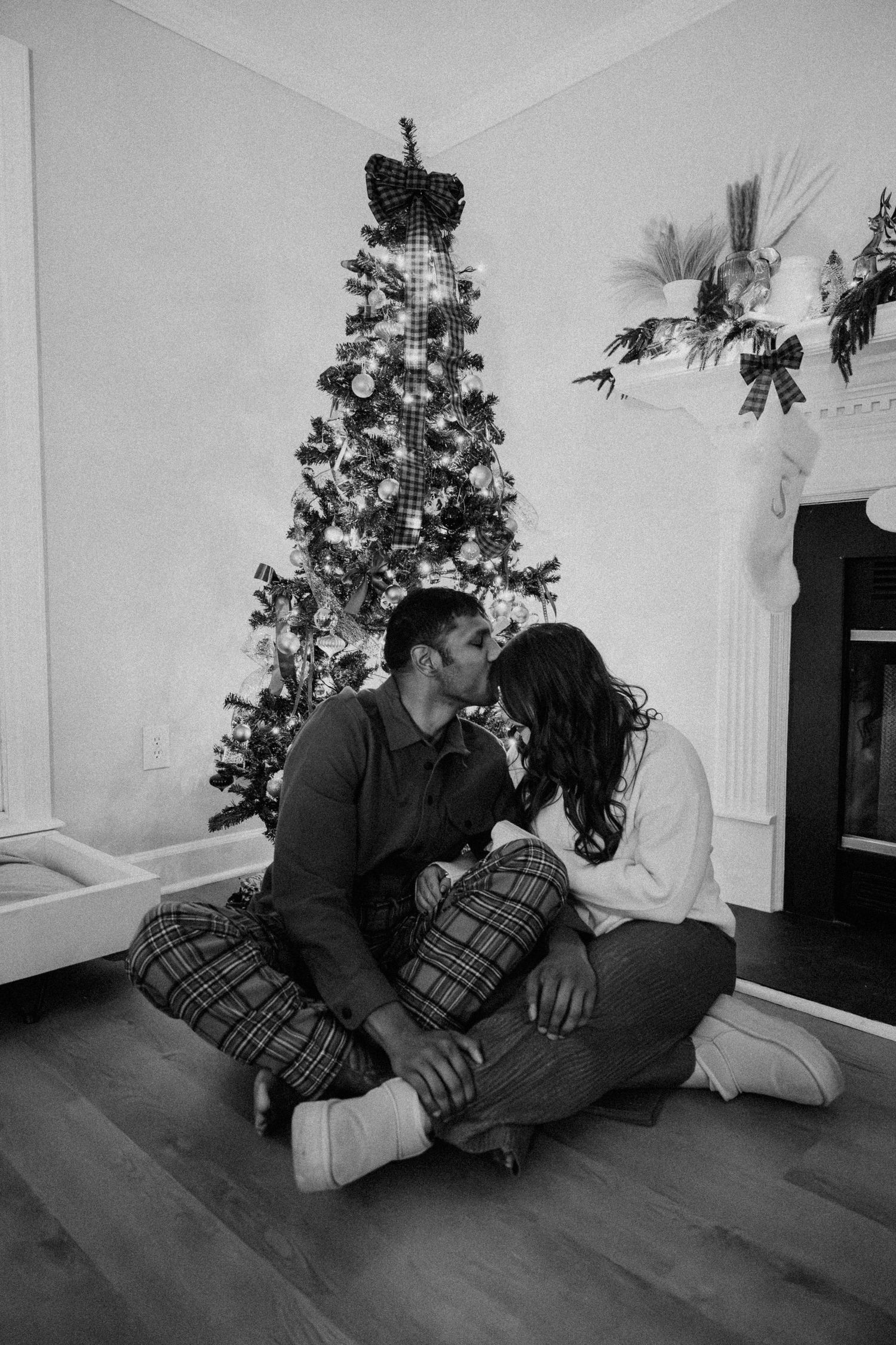 Black and white photo of a couple kissing in front of a decorated Christmas tree by a fireplace on Christmas morning.