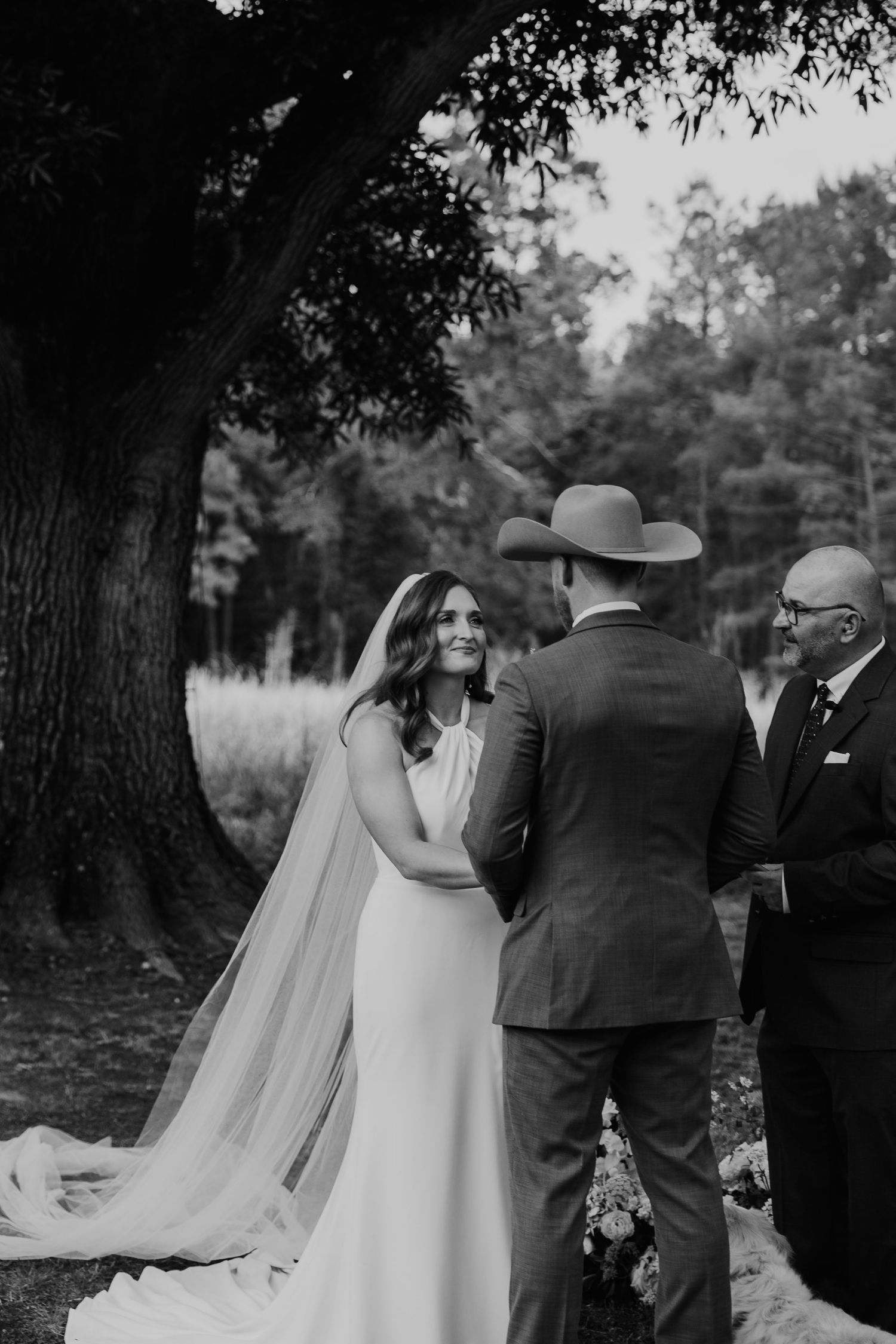 Black and white wedding ceremony photo of bride in veil and groom in cowboy hat exchanging vows under large tree.
