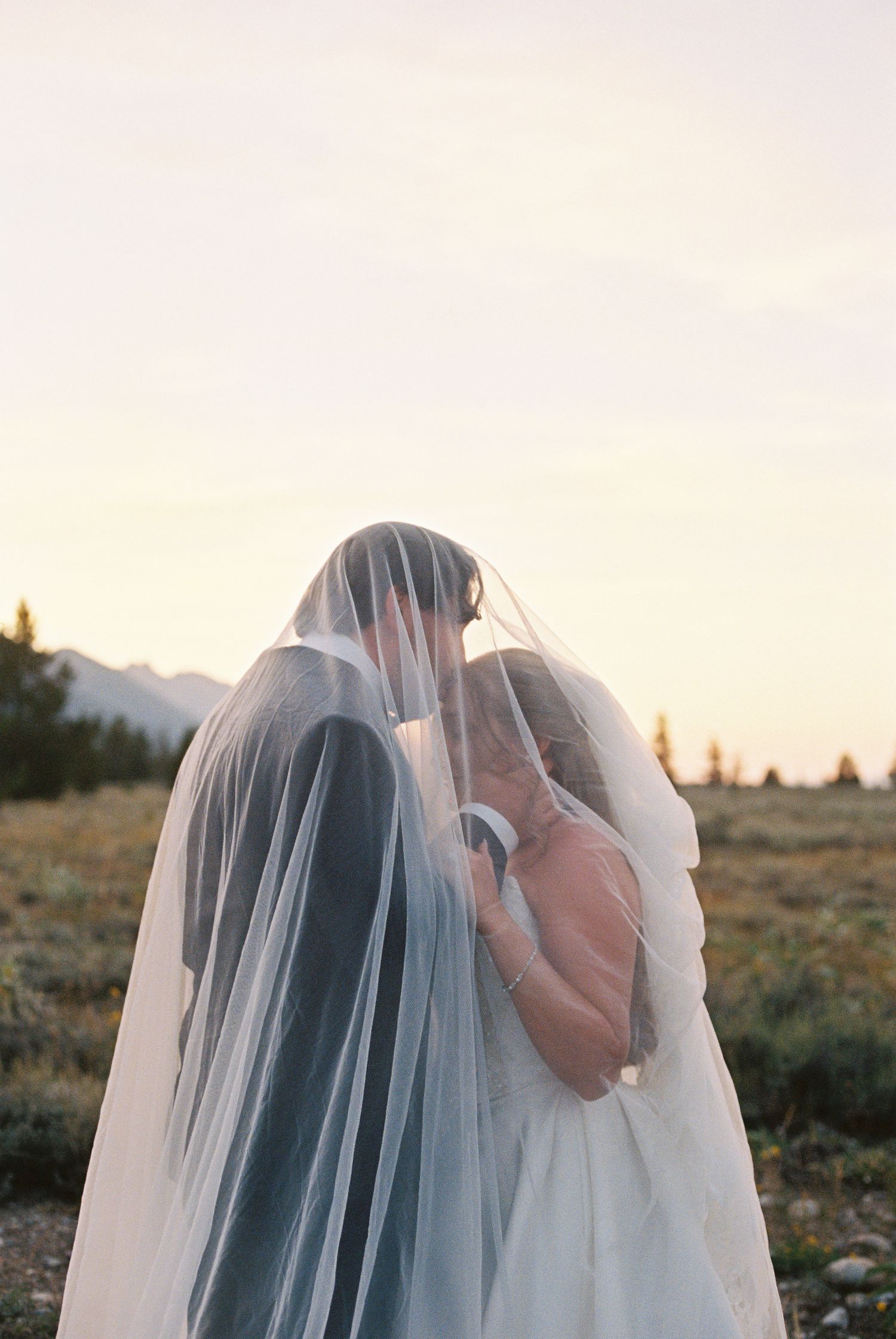 Bride and groom sharing a kiss under a flowing wedding veil at sunset with mountain landscape backdrop.