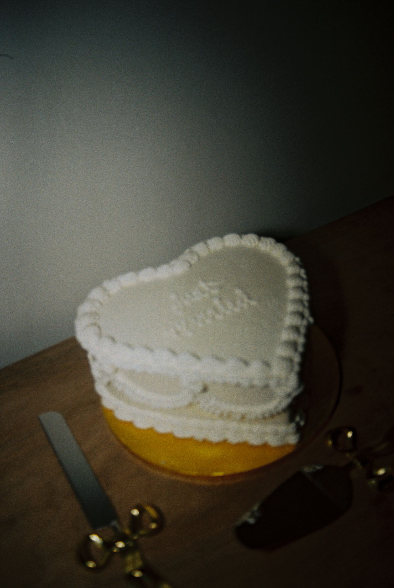 White heart-shaped wedding cake with piped frosting border and script writing, displayed on wooden table with gold cake servers.