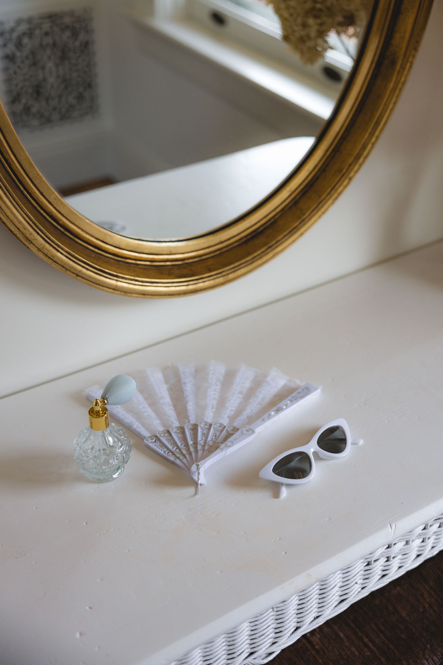 Gold oval mirror above a white vanity with a glass perfume bottle, white lace fan, and cat-eye sunglasses.