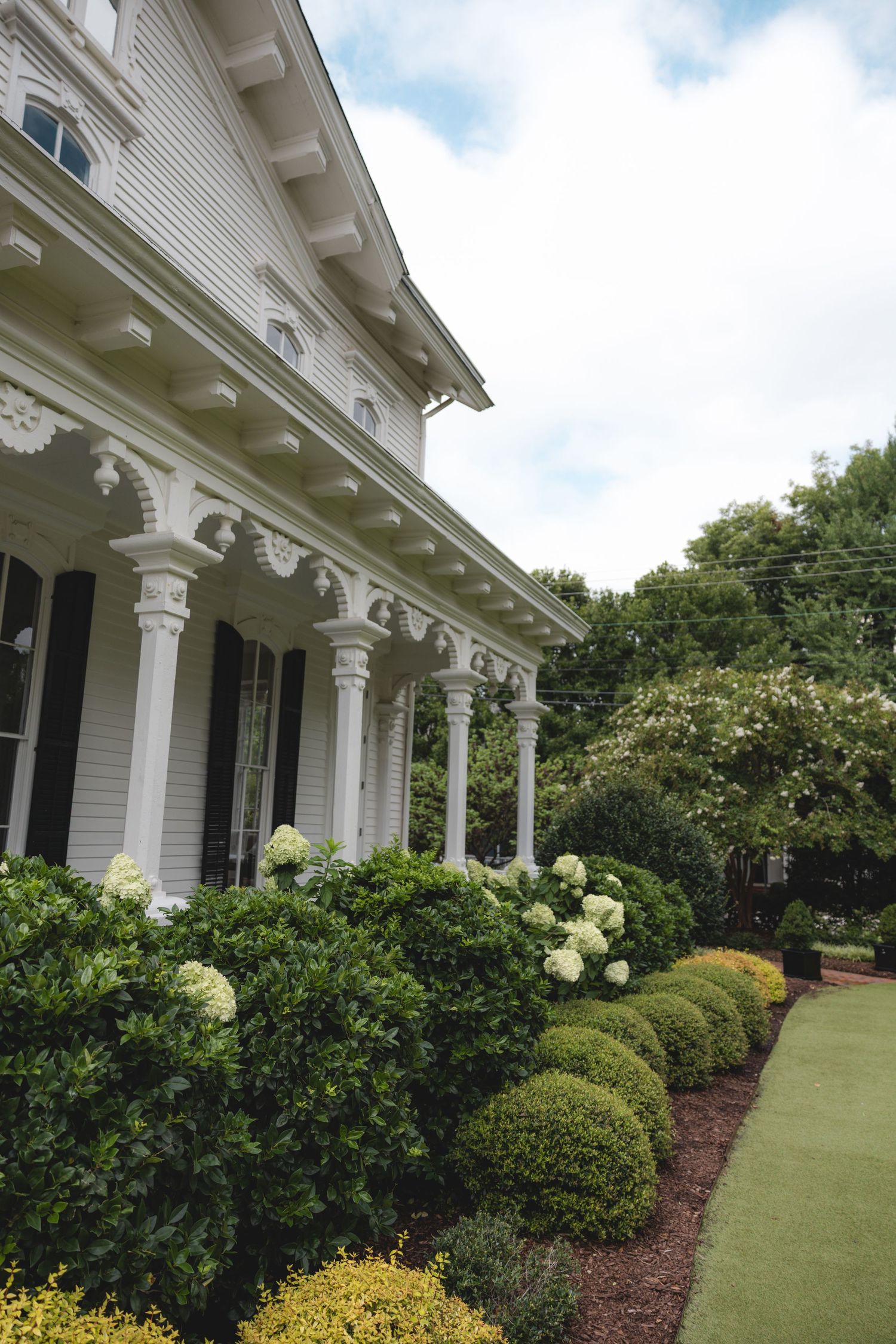 White colonial mansion with ornate columns, black shutters, and lush green landscaped gardens with hydrangeas.