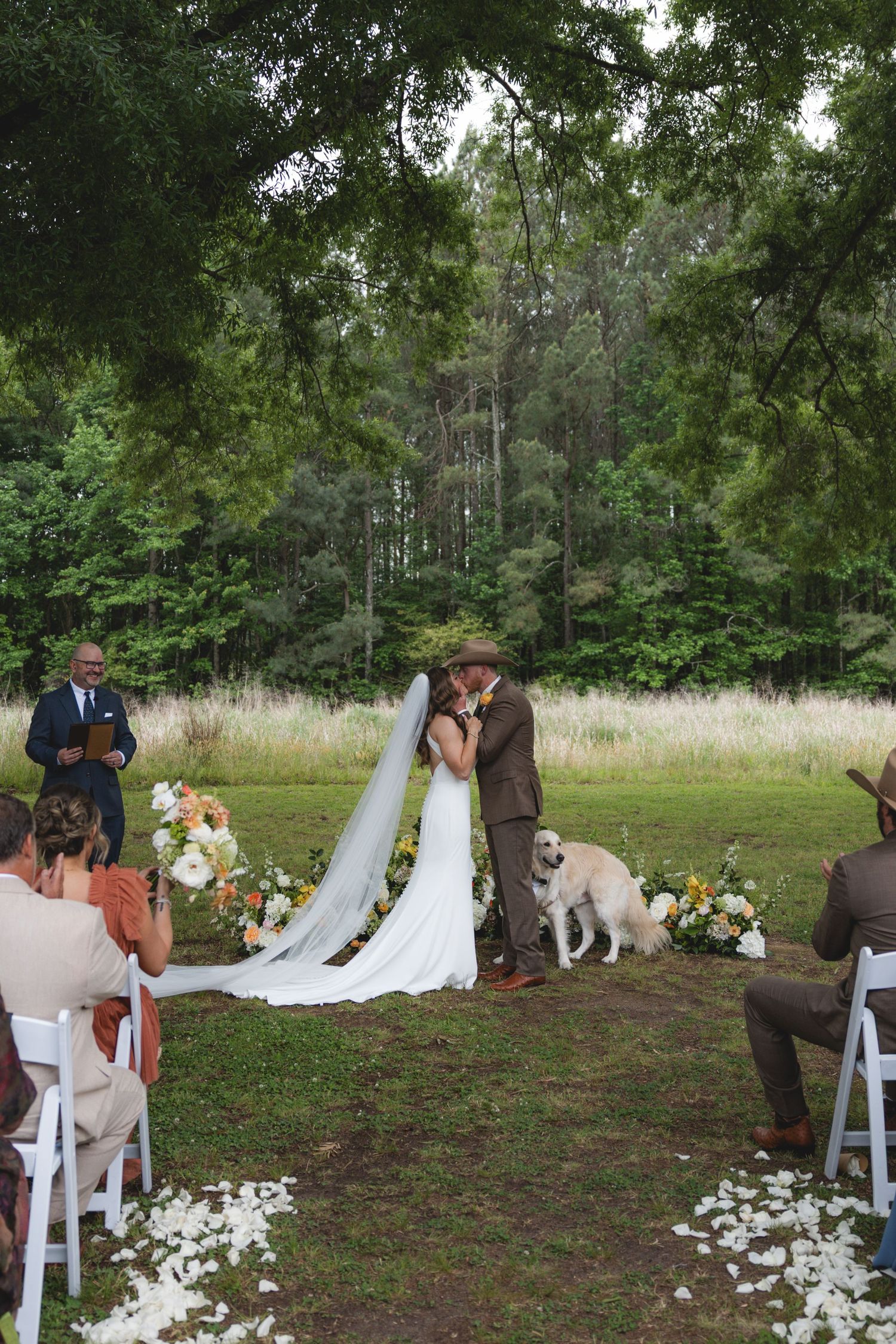 Couple shares first kiss at outdoor wedding ceremony with white chairs, floral arrangements, and a dog beside them.