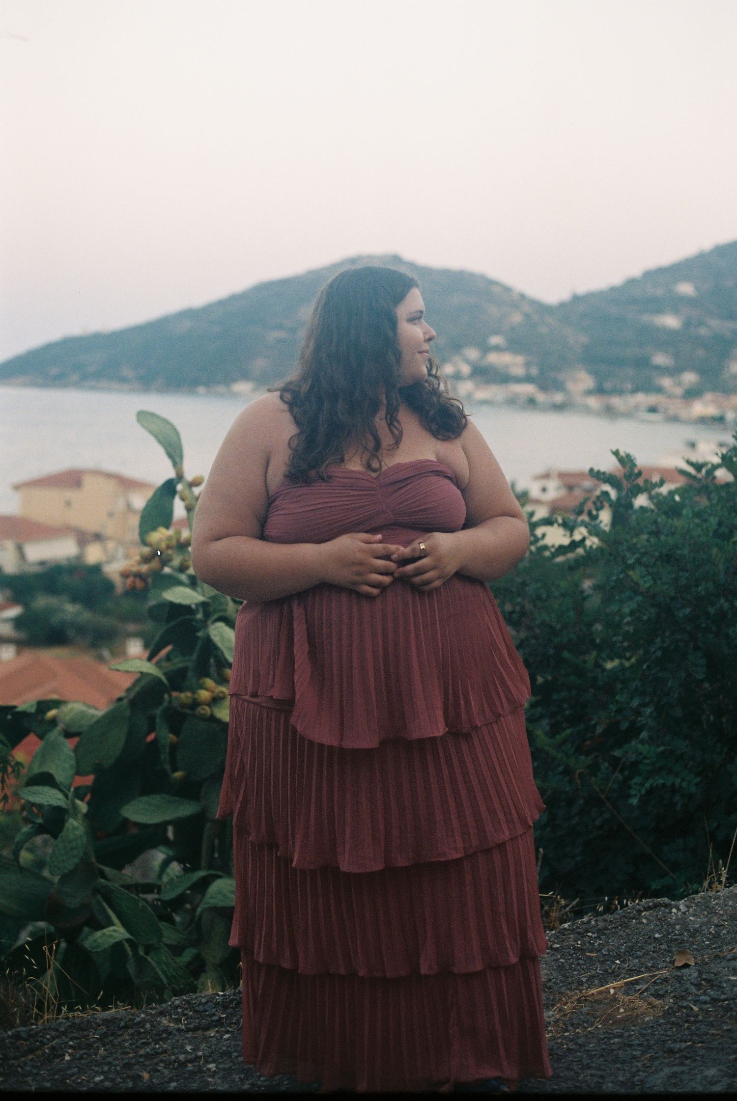 Plus-size woman in a layered burgundy strapless gown poses with a scenic coastal village backdrop at dusk.