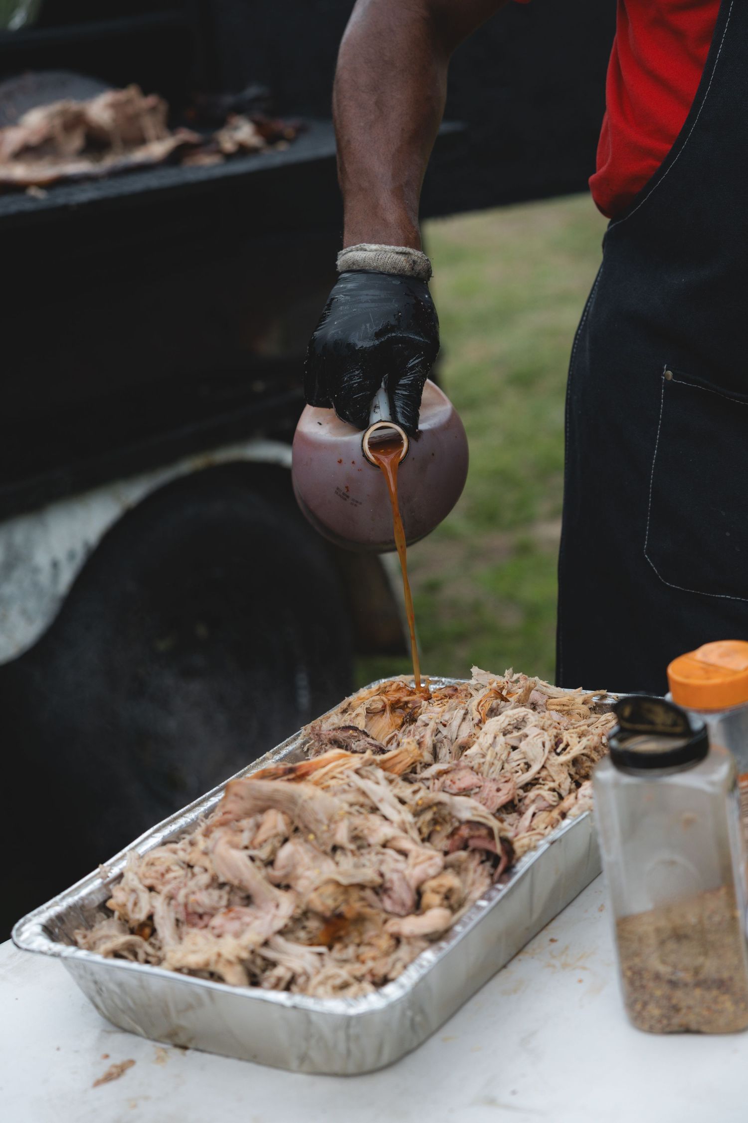 Caterer pouring BBQ sauce over pulled pork in a foil pan outdoors.