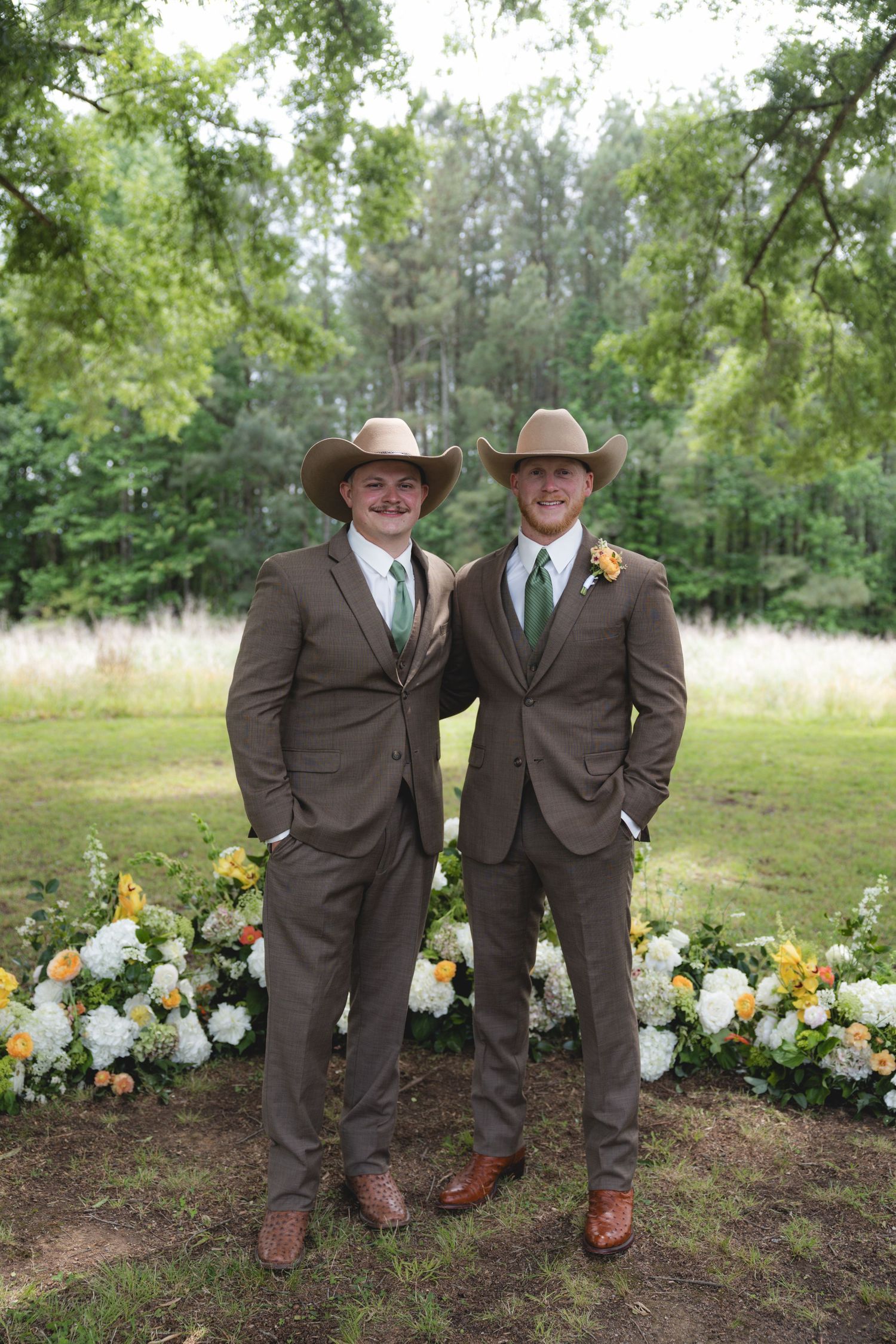 Two groomsmen in matching brown suits and cowboy hats pose before floral arrangement outdoors.