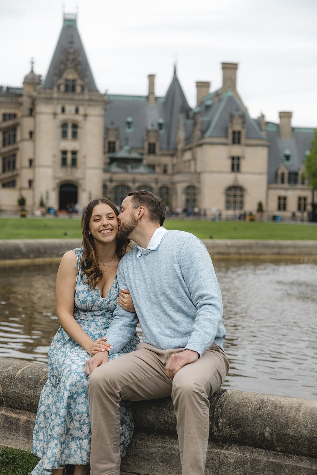 Couple sitting by a fountain at Biltmore Estate, man kissing woman on cheek as she smiles, wearing floral dress.