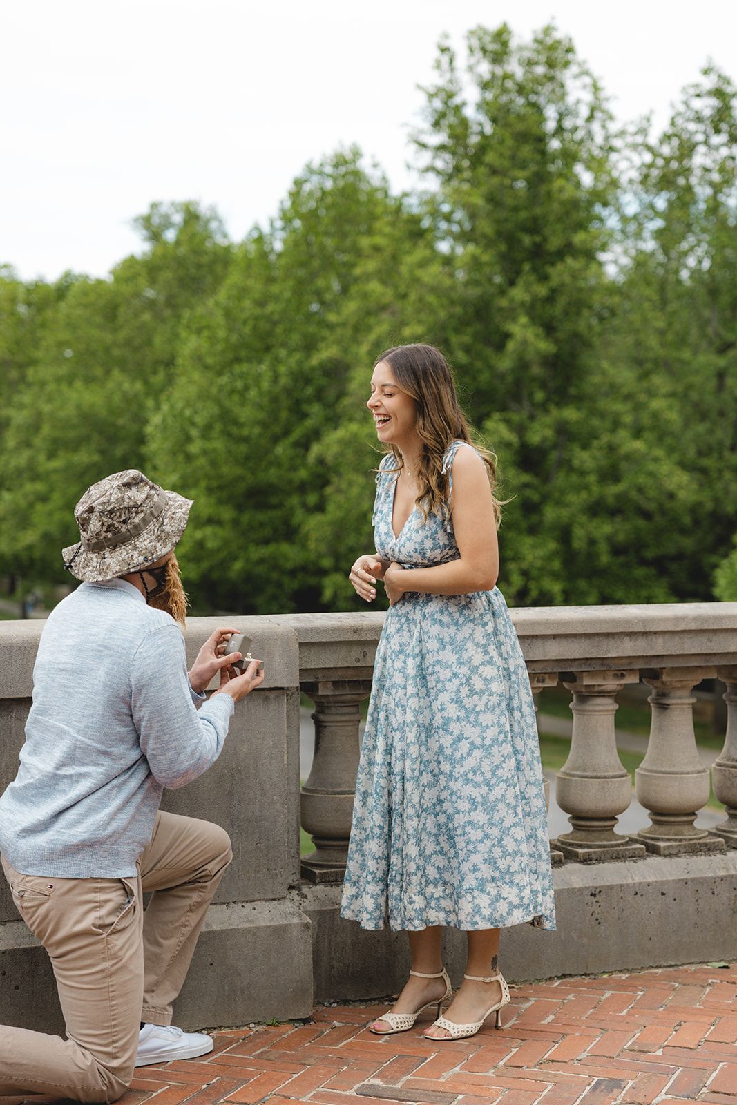 Man kneeling to propose to a laughing woman in a floral dress on a stone terrace in a lush green park.