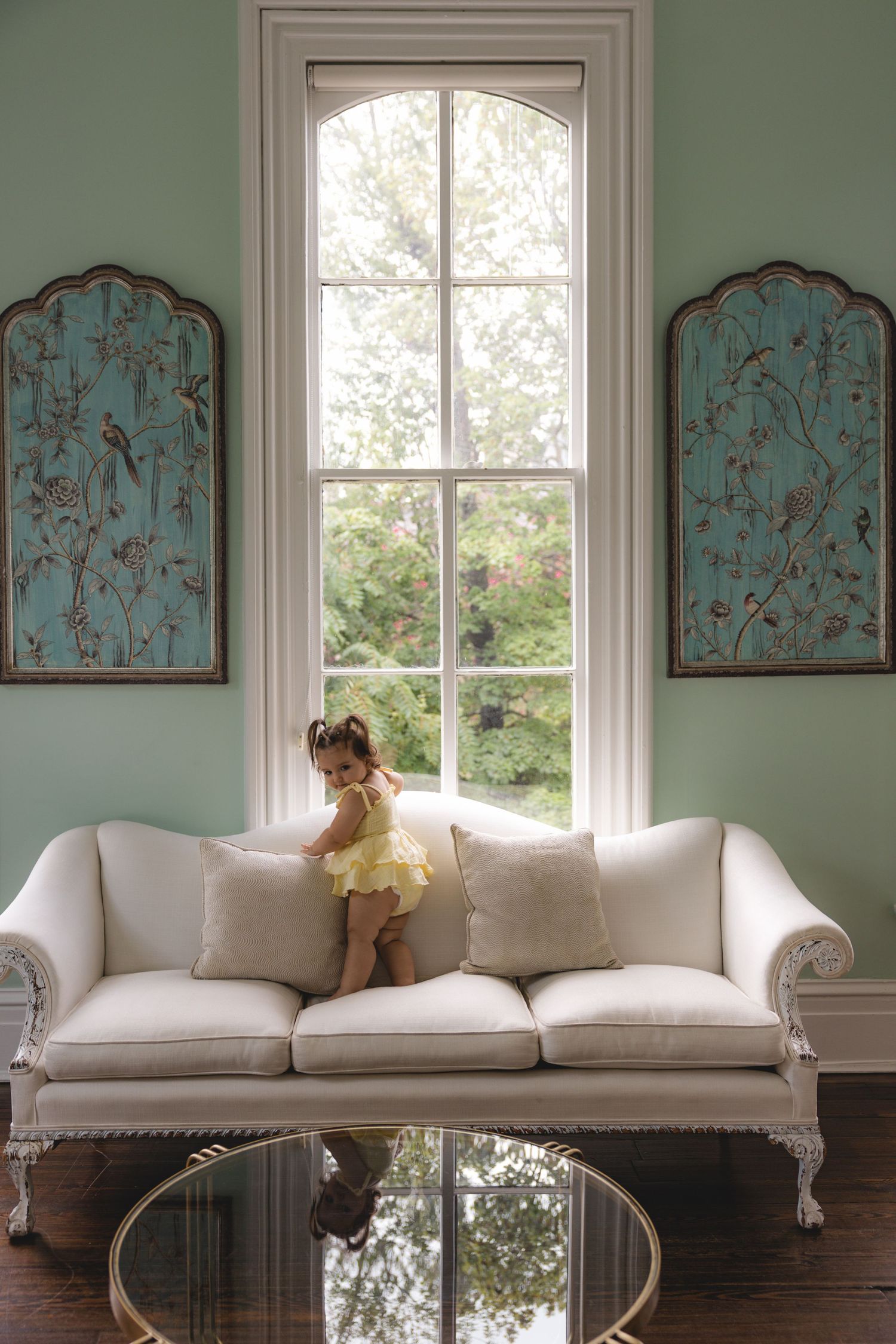 Young girl in yellow dress standing on white ornate sofa by tall arched window with blue floral wall panels.