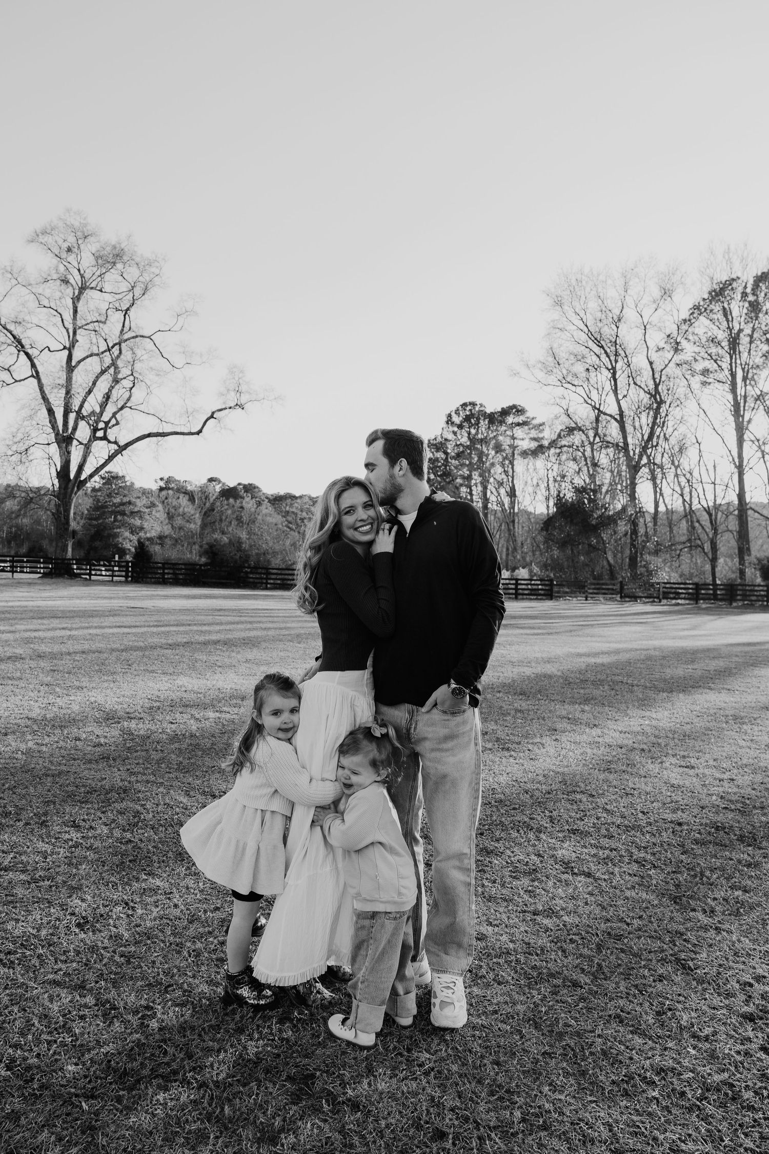 Black and white family photo of couple with two young children standing in grassy field with trees in background.