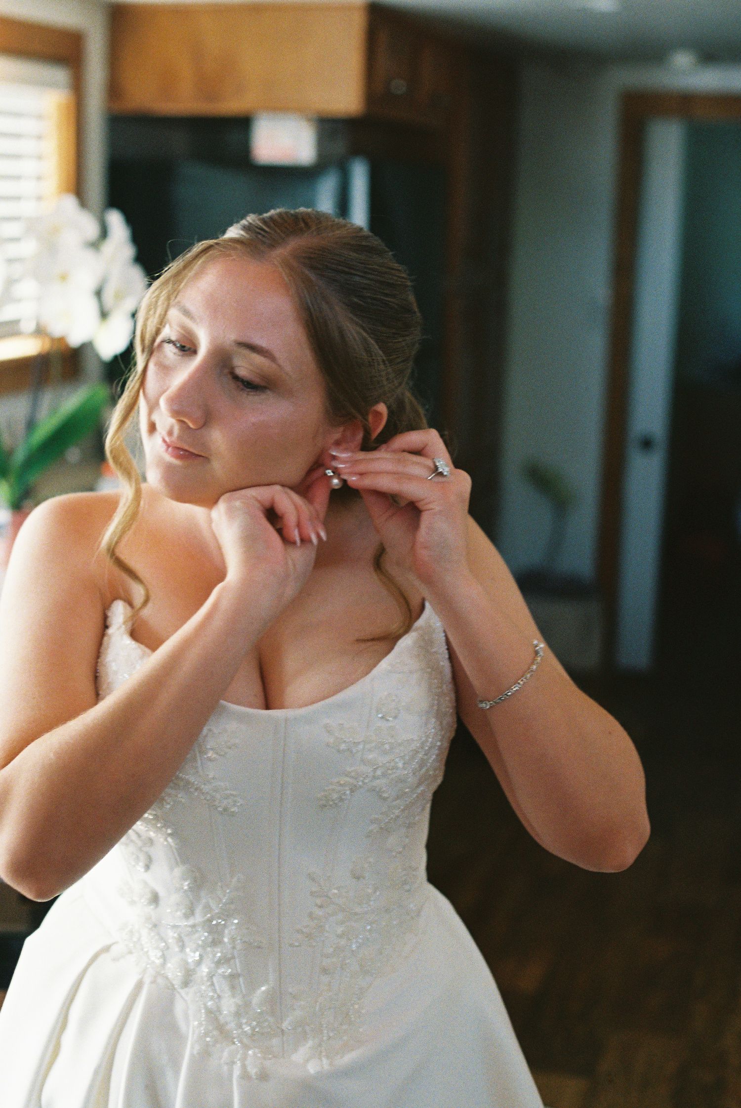 Bride in strapless lace wedding gown putting on pearl earrings while getting ready for her wedding day.