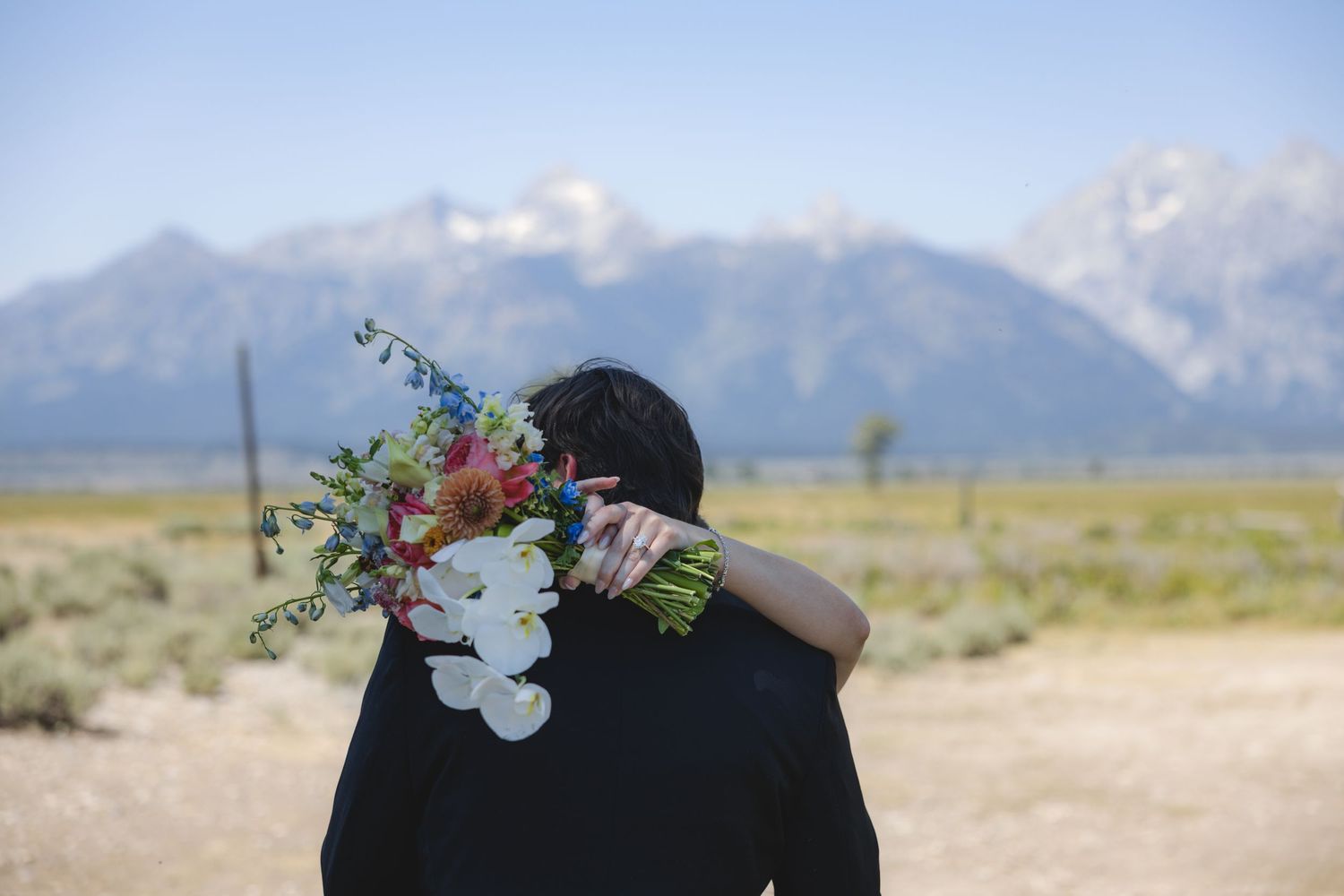Couple embracing with wildflower bouquet, scenic Grand Teton mountain range in background on clear day.