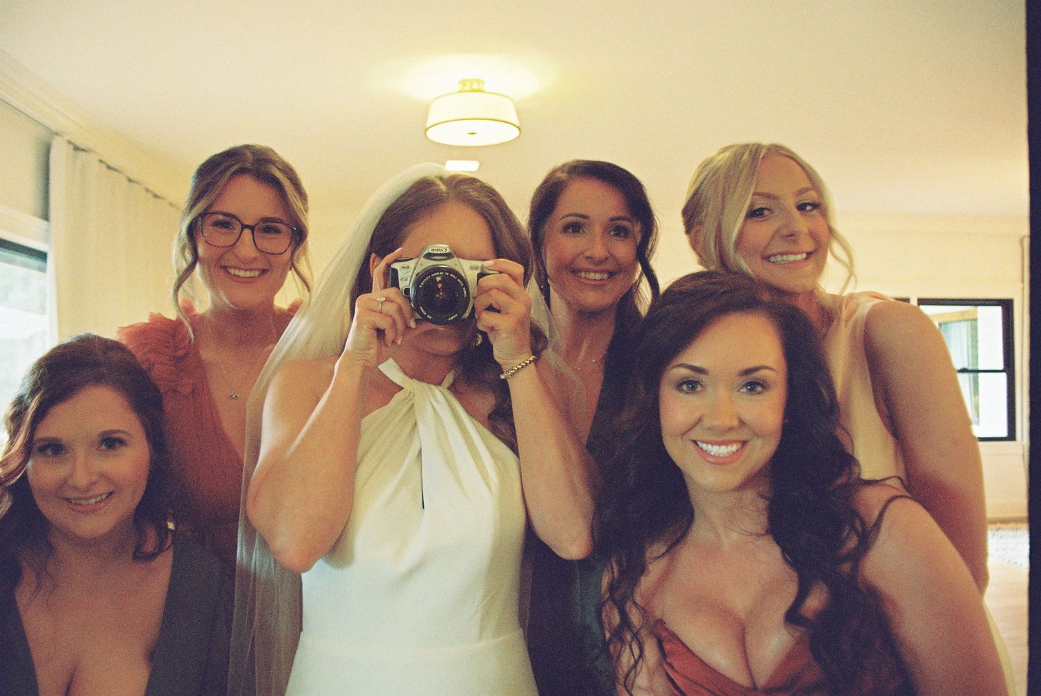 Bride in white dress takes mirror selfie with five bridesmaids in a bright room before wedding ceremony.