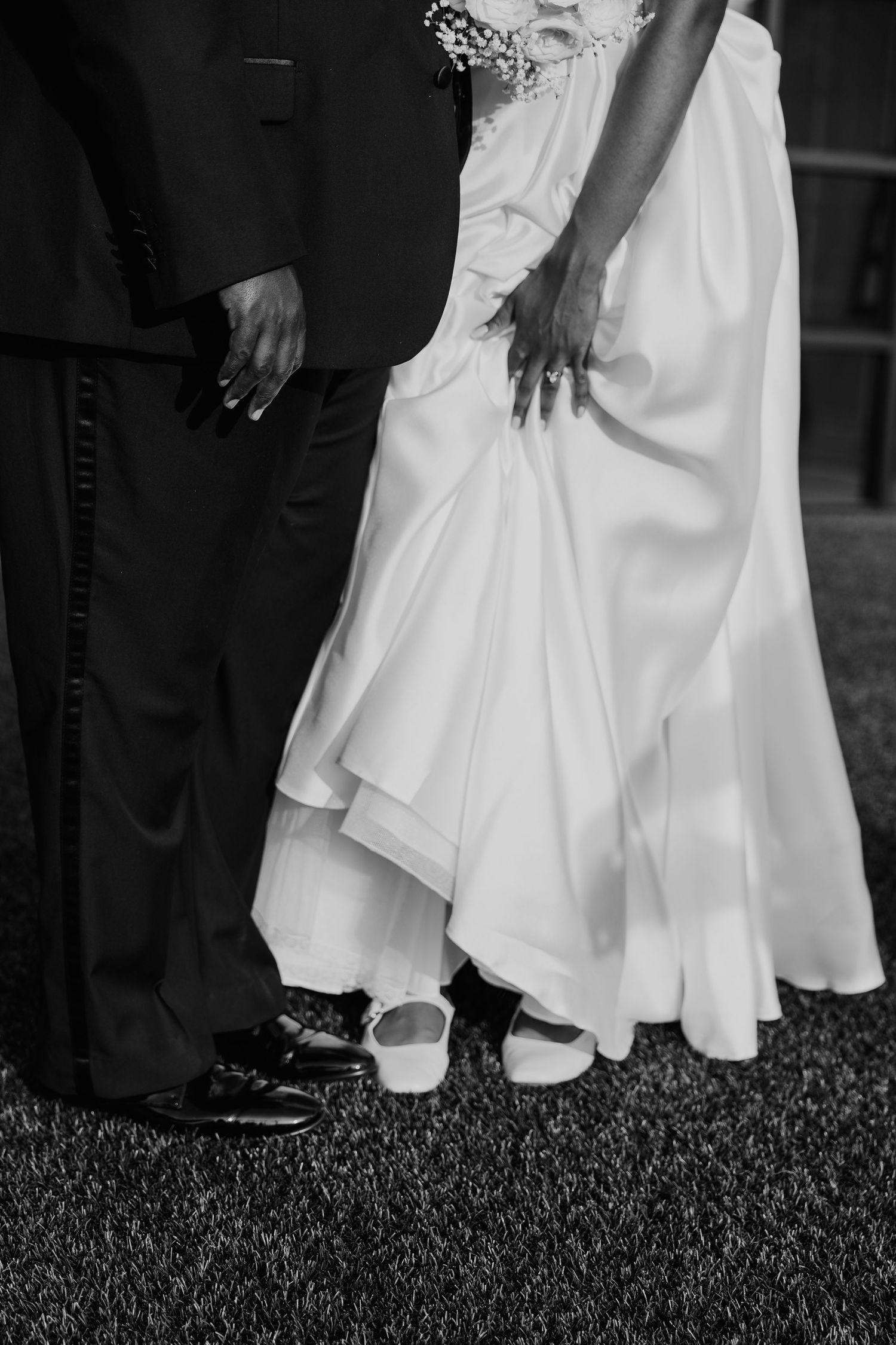 Black and white wedding photo showing bride in white gown and groom in dark suit standing on grass, hands touching.