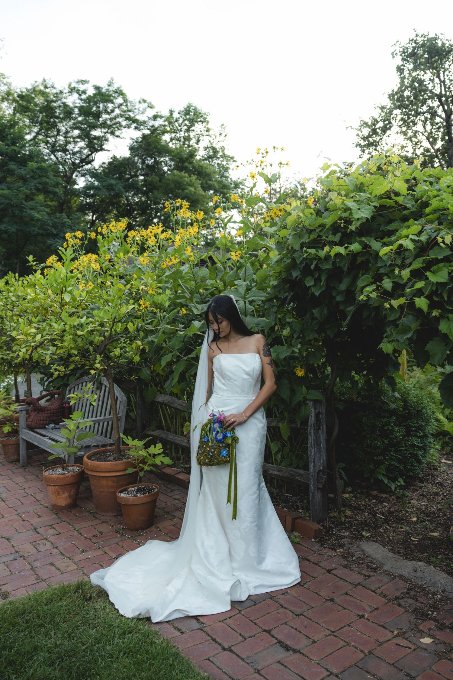 Bride in white strapless gown with veil holds colorful bouquet in lush garden with brick pathway.