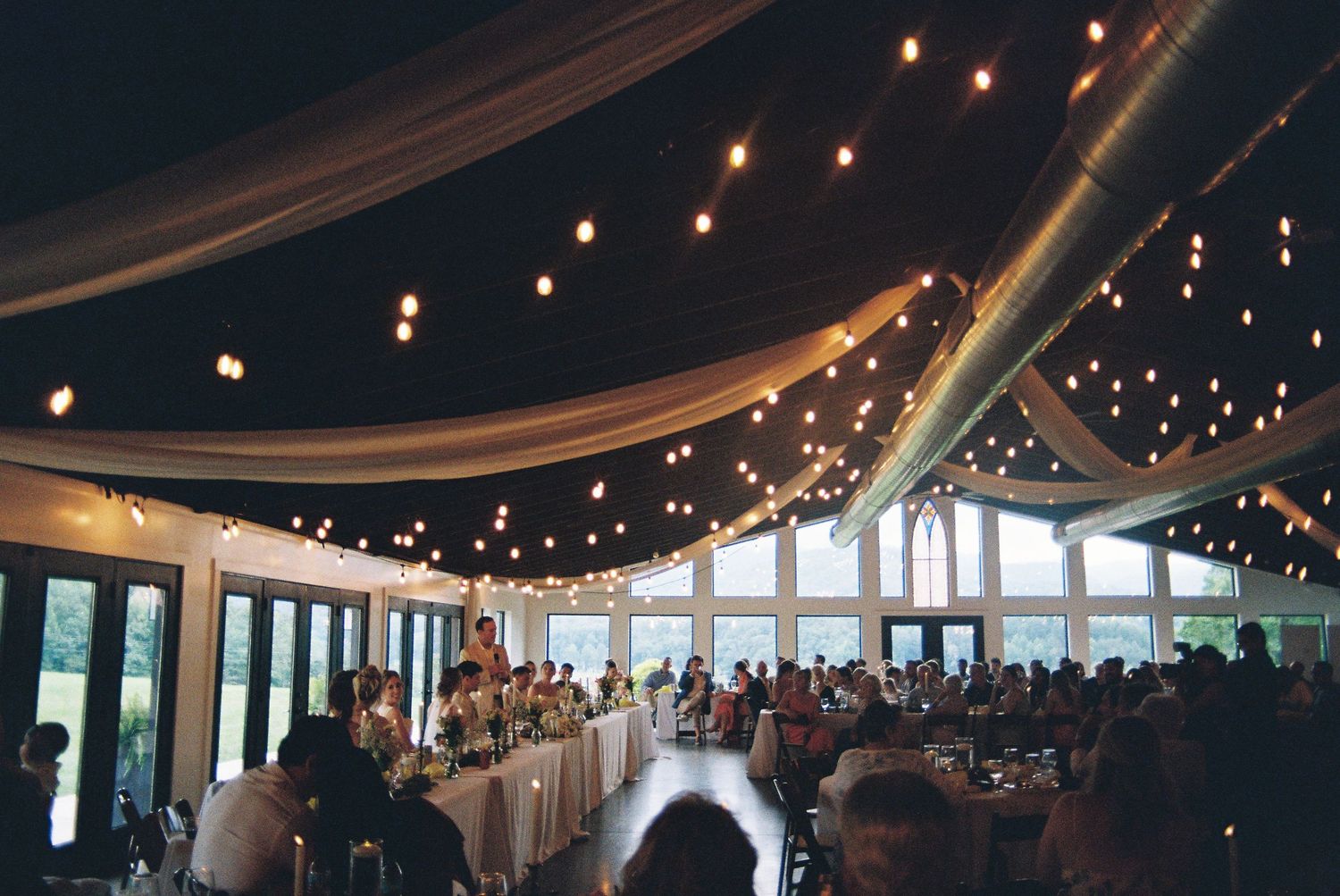 Elegant wedding reception venue with string lights and draped fabric ceiling, guests dining at decorated tables.