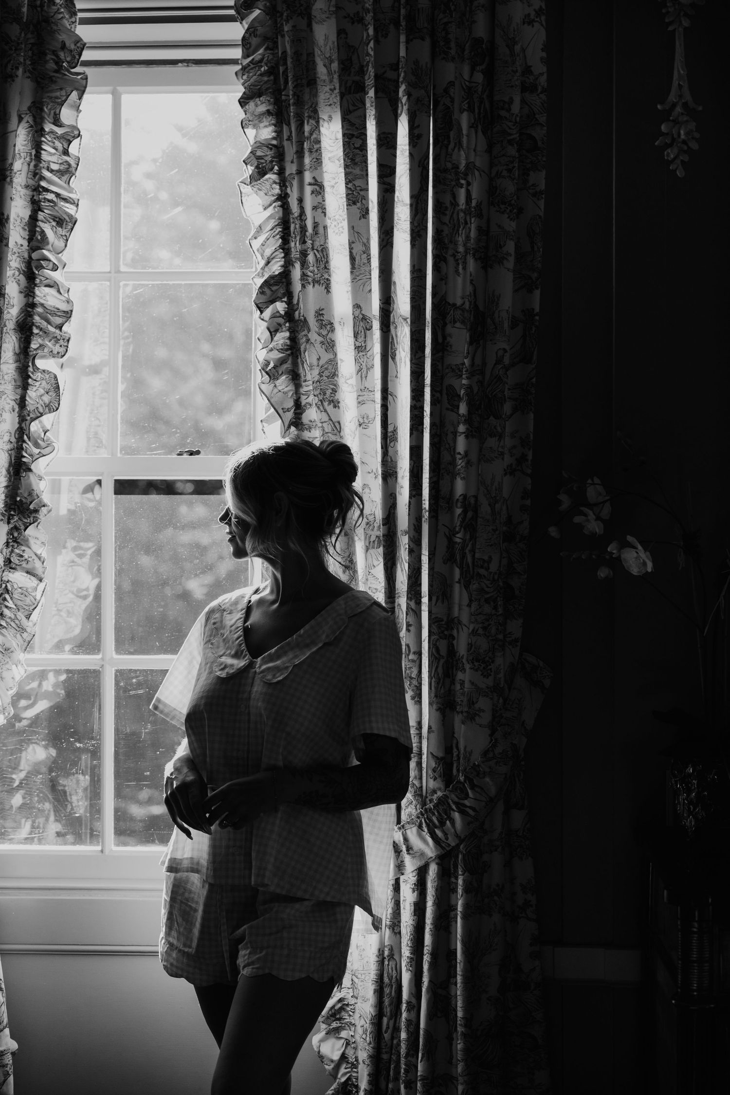Black and white photo of a bride in pajamas gazing out a window with floral curtains on her wedding morning.