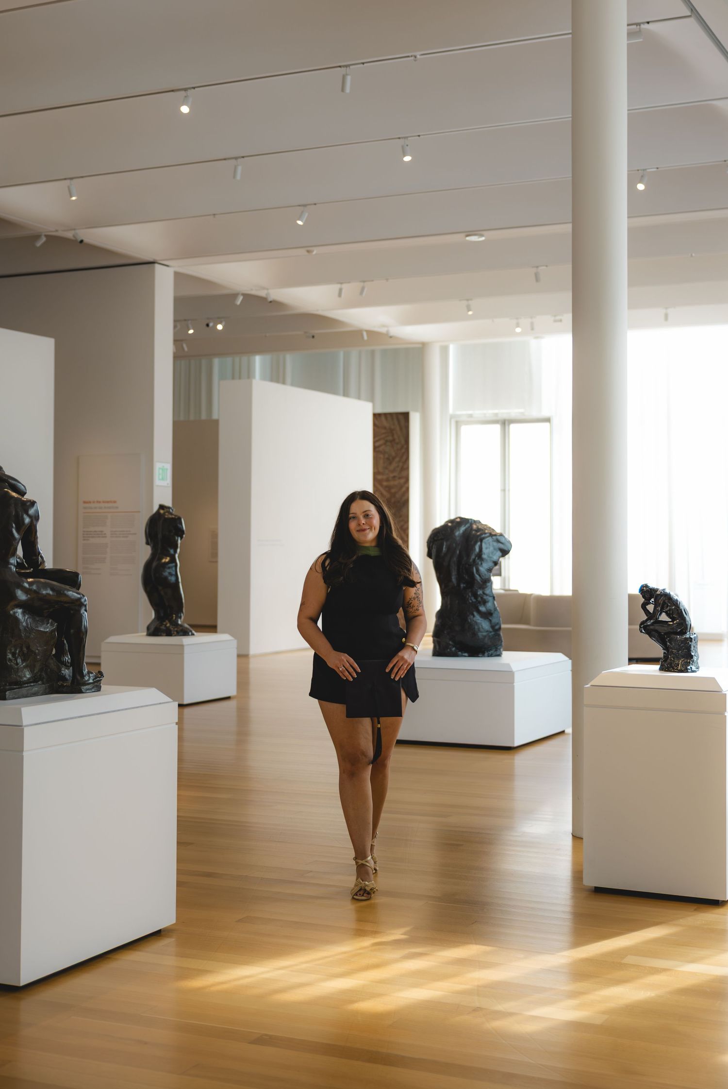 Woman in black dress walking through a modern art museum gallery with bronze sculptures on white pedestals.