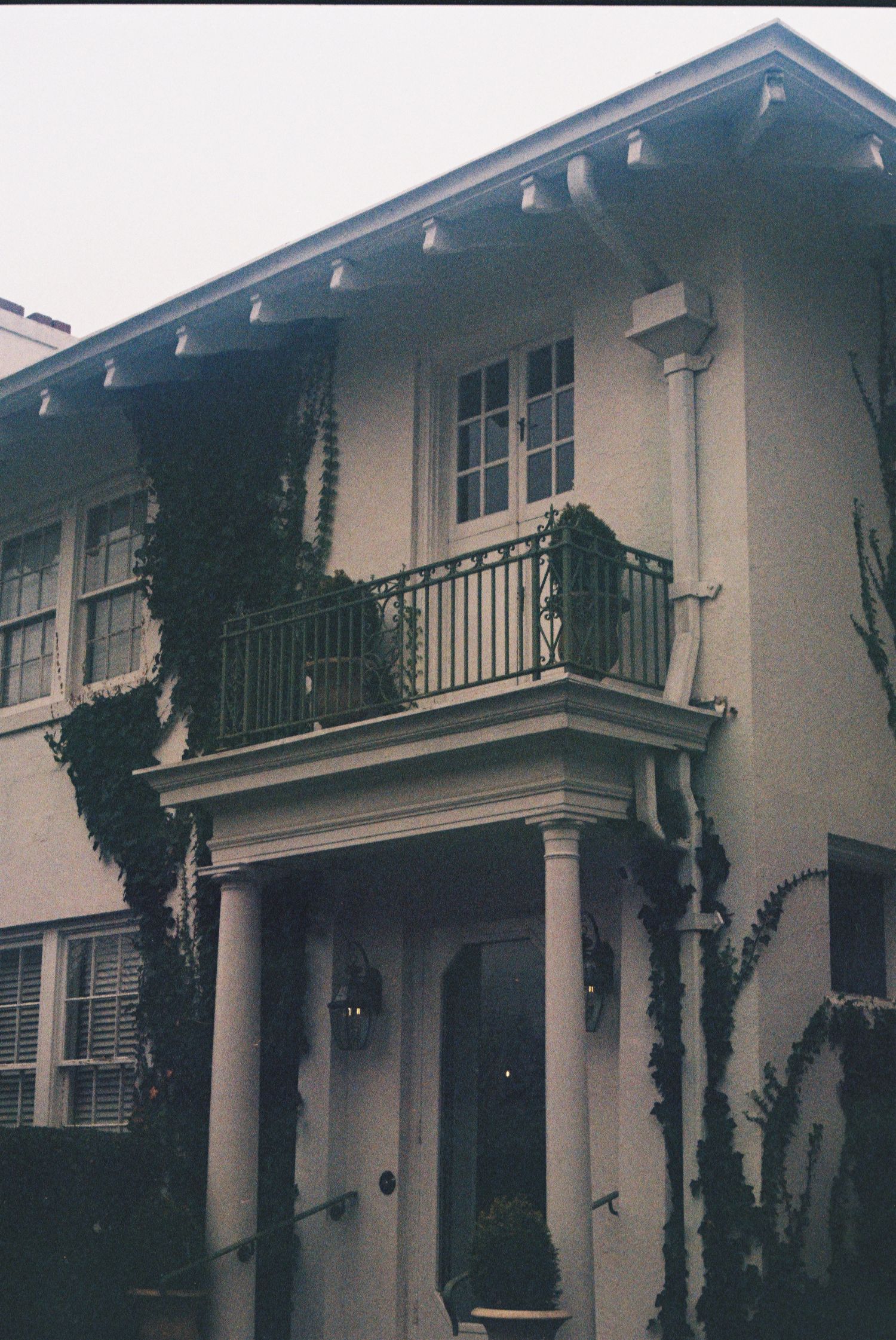 The image shows a historic-looking two-story building with a balcony, surrounded by lush greenery.