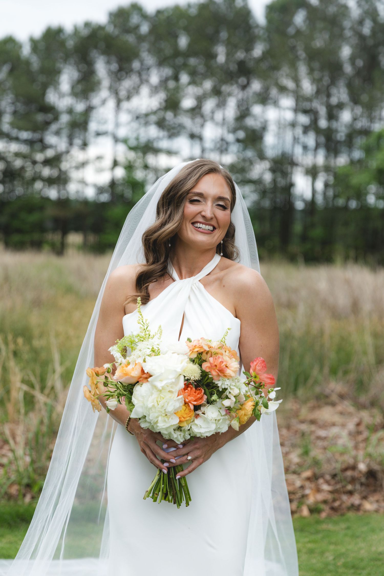 Smiling bride in white halter gown and veil holds peach and white bouquet in front of pine tree backdrop.