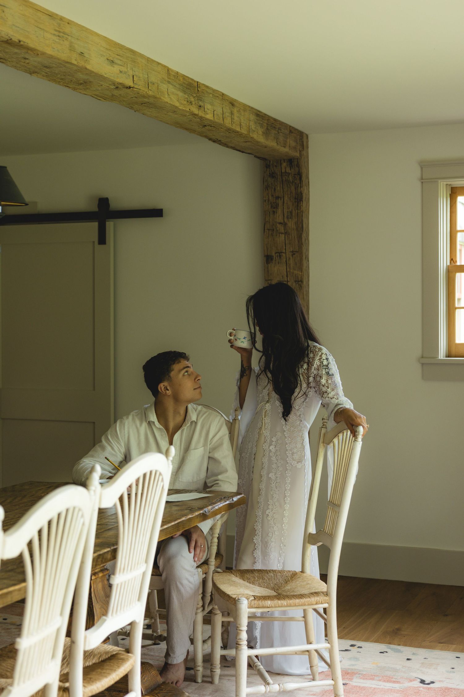 Couple in a rustic dining room, man seated at table looking up at woman in white lace dress holding a cup.