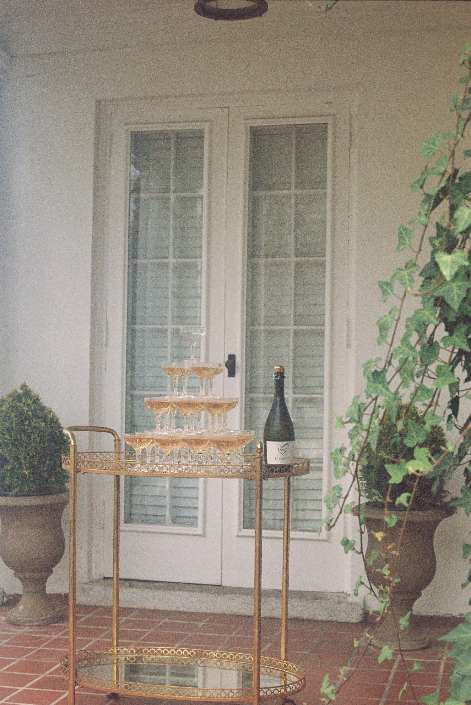 Gold bar cart with champagne tower and bottle beside French doors, flanked by potted boxwood and ivy on a brick patio.