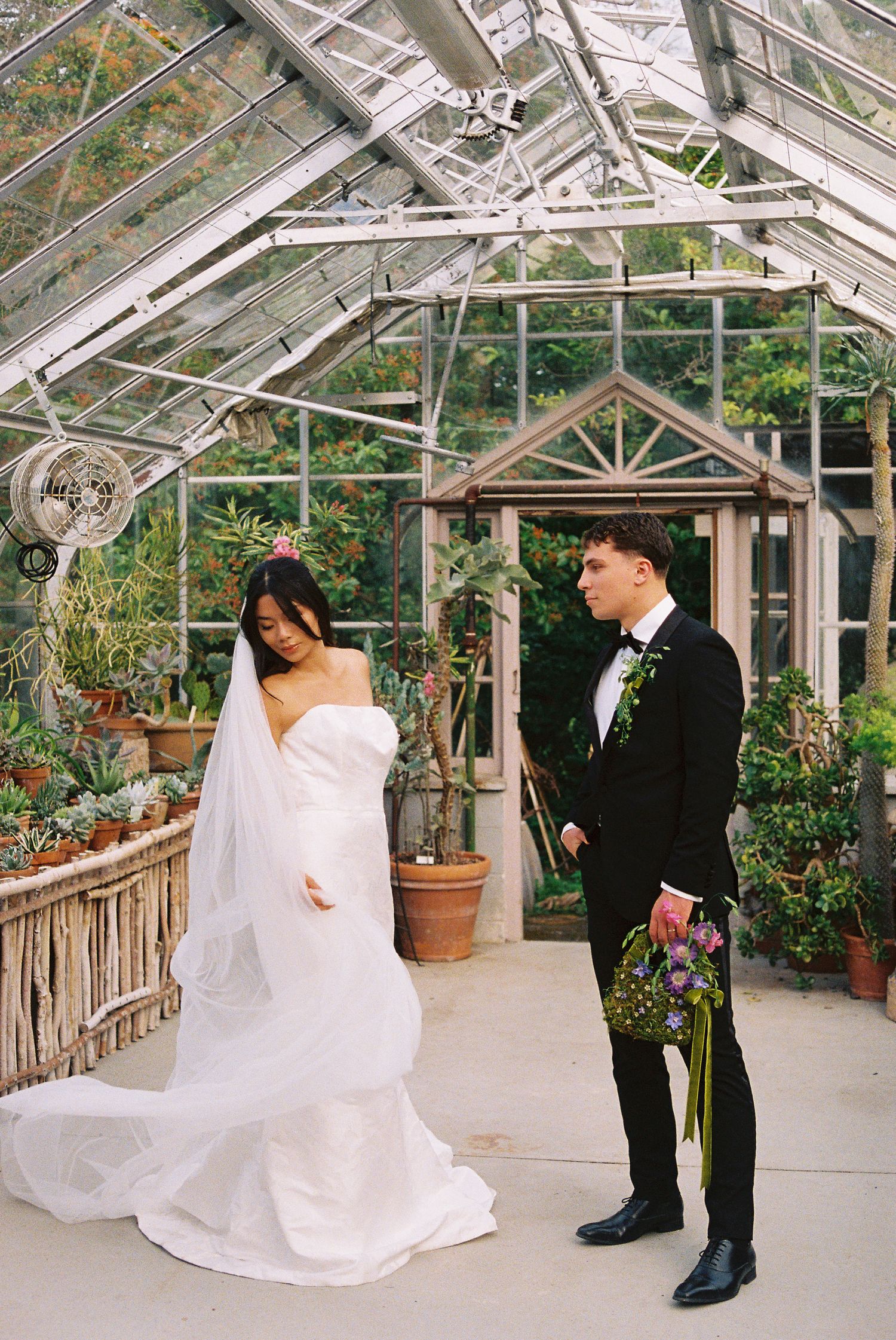 Bride in white gown and groom in black tuxedo share a romantic moment inside a lush greenhouse venue.