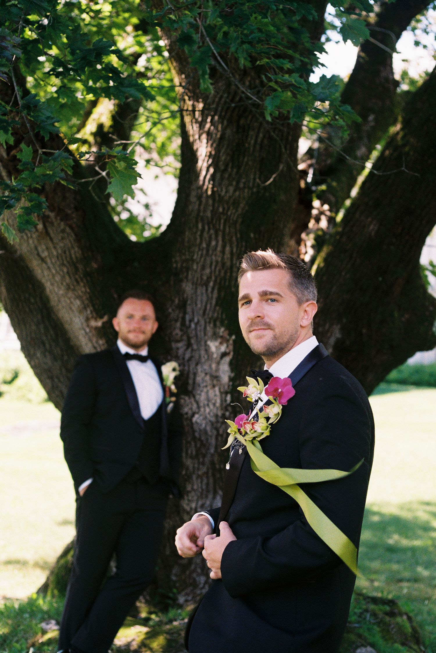 Two grooms in black tuxedos with floral boutonnieres pose near a large tree outdoors at a wedding.