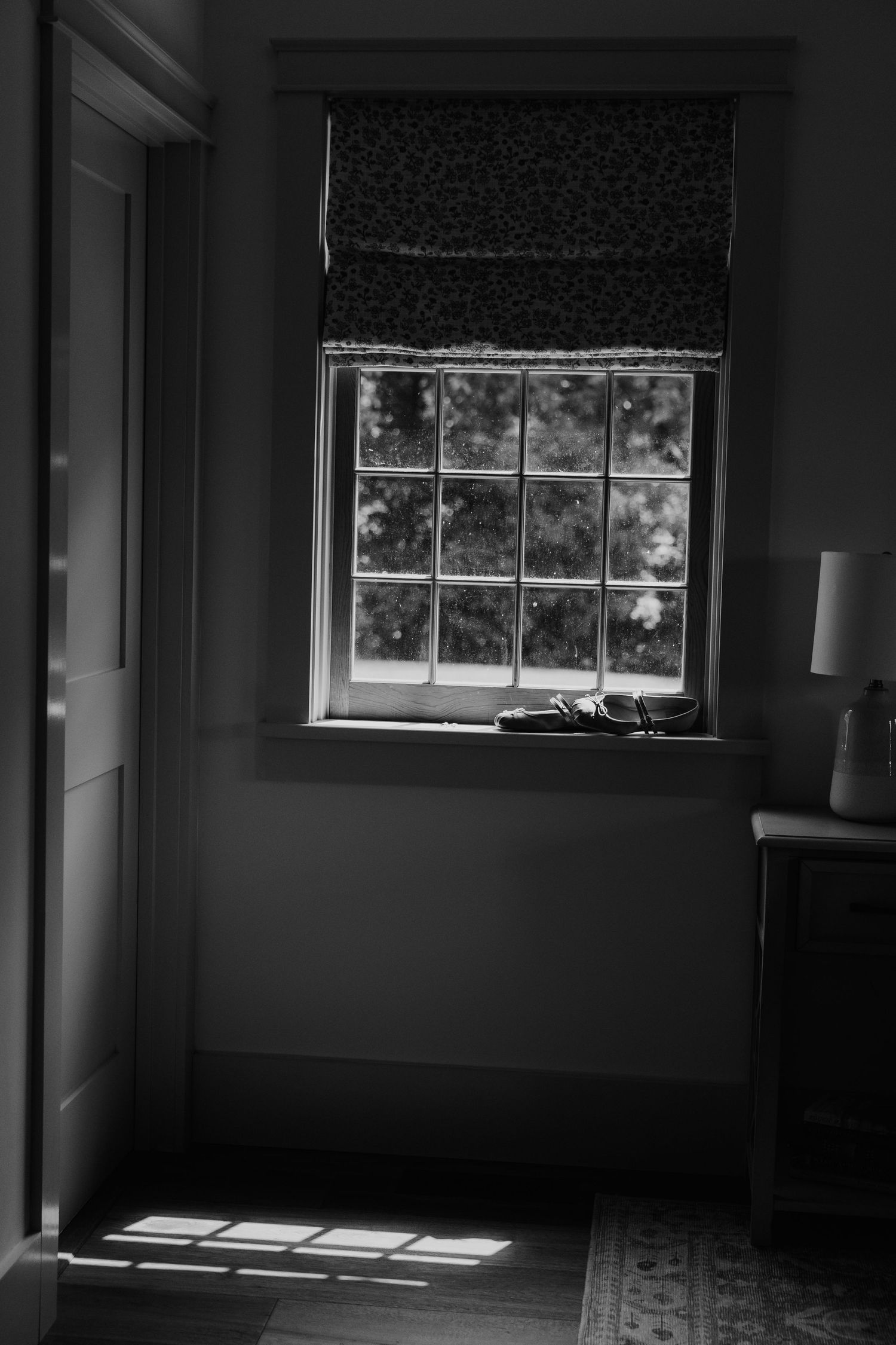 Black and white photo of sunlight streaming through a multi-pane window with a Roman shade in a dimly lit room.